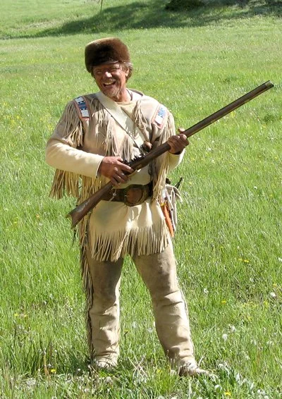 Mountain Man Mike Guli, in buckskin outfit, holding his flintlock rifle, on a grassy field.