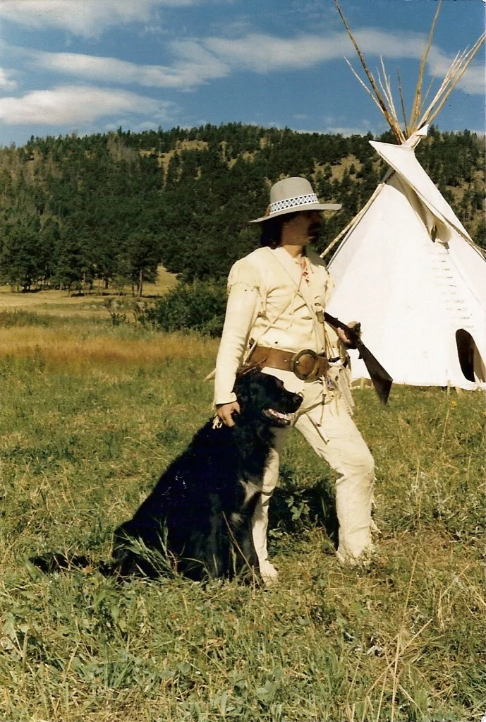 A person dressed in Old West style clothing, holding a rifle and standing next to a black and white dog, in front of a teepee with a forested hillside in the background.