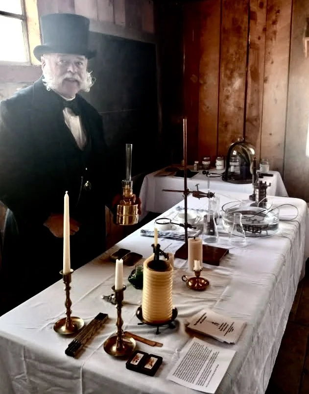 A man dressed in period clothing, including a top hat and suit, standing next to a historical science or alchemy display on a table with scientific instruments, candles, and documents inside a wooden-paneled room.