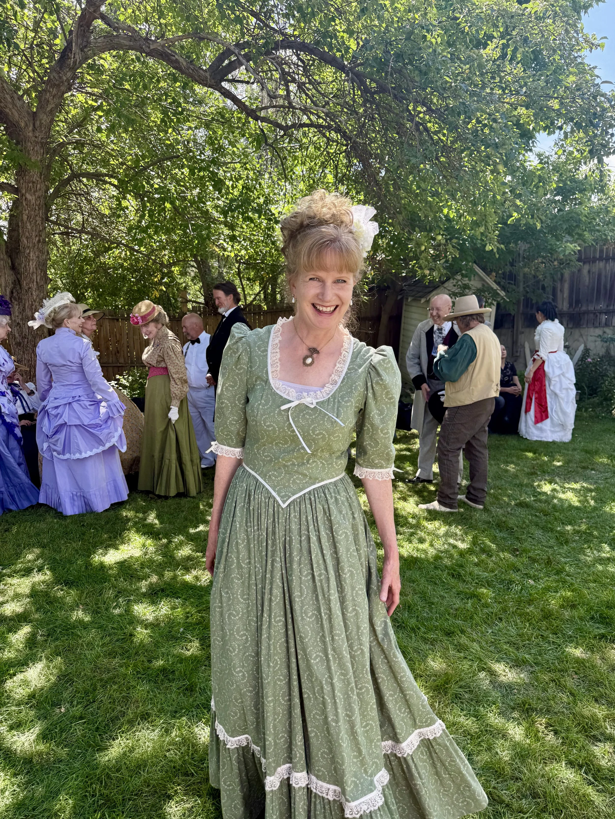 Woman in historical green dress smiling outdoors at a gathering with other people dressed in vintage clothing under a large tree.