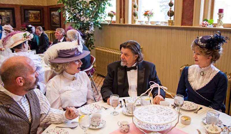 People dressed in vintage clothing, sitting at a decorated dining table during a tea party in a well-lit room.