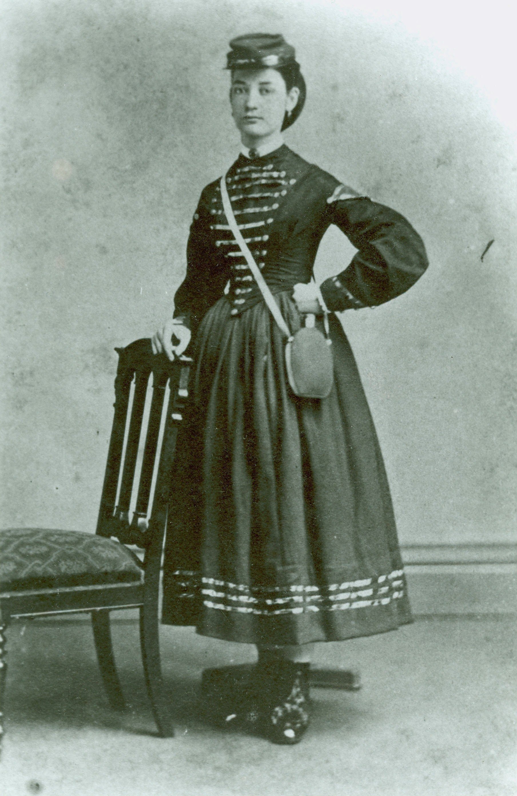 Black and white photograph of a woman standing with one hand on her hip, dressed in a historical military-style uniform, wearing a hat, with a small canteen hanging from her shoulder, standing next to a chair.