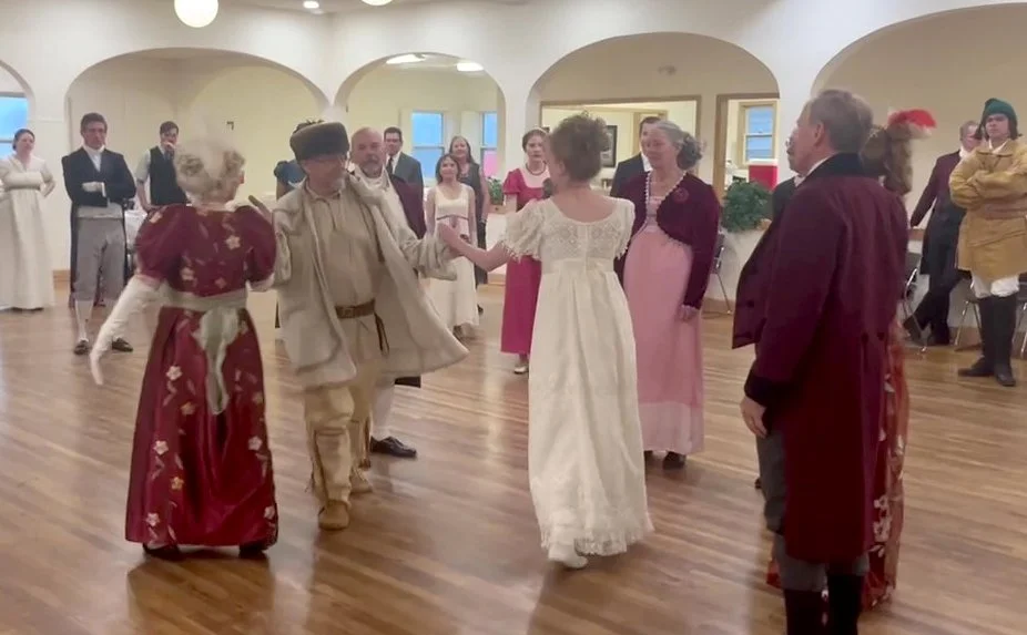 People dressed in Lewis and Clark or Regency costumes dancing in a room with wooden floors and white walls.