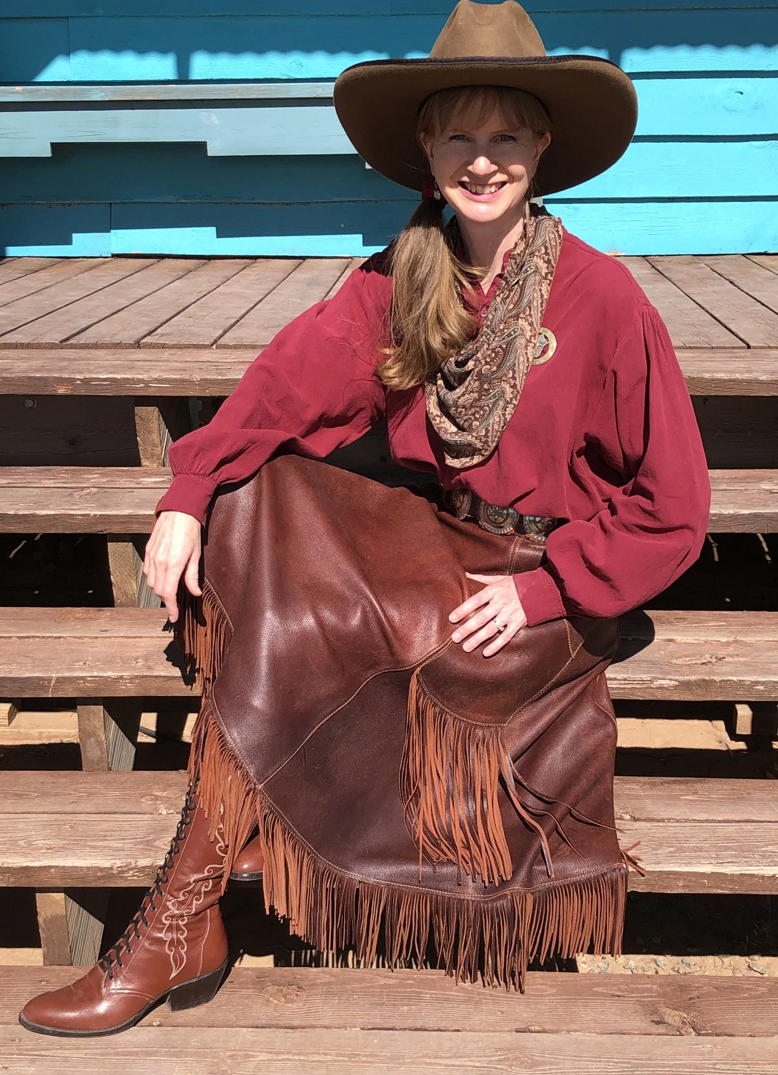 Sharon Guli in an Old West Cowgirl outfit, with wide-brimmed hat, neckerchief, red blouse, leather fringed skirt, and lace-up boots, sitting on steps in front of a building.