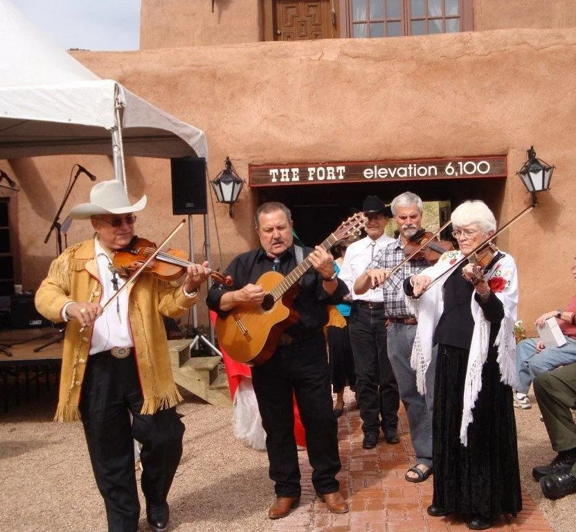 Group of five musicians playing instruments outdoors near a building with a sign that reads 'The Fort elevation 6,100'. They are dressed in Western or rustic style clothing, including cowboy hats, vests, and shawls. One man is playing a violin, another a guitar, another a violin, and a woman is playing a violin.