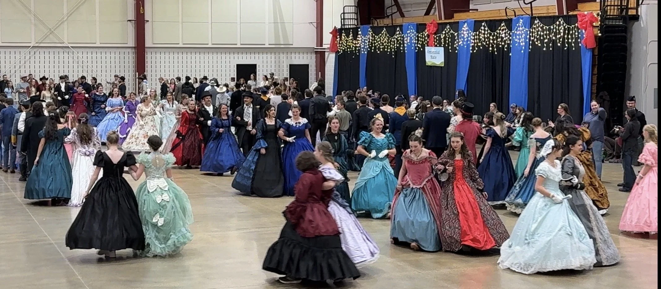 People dressed in Victorian costumes dancing in a large indoor hall decorated with string lights and banners for a historical ball.