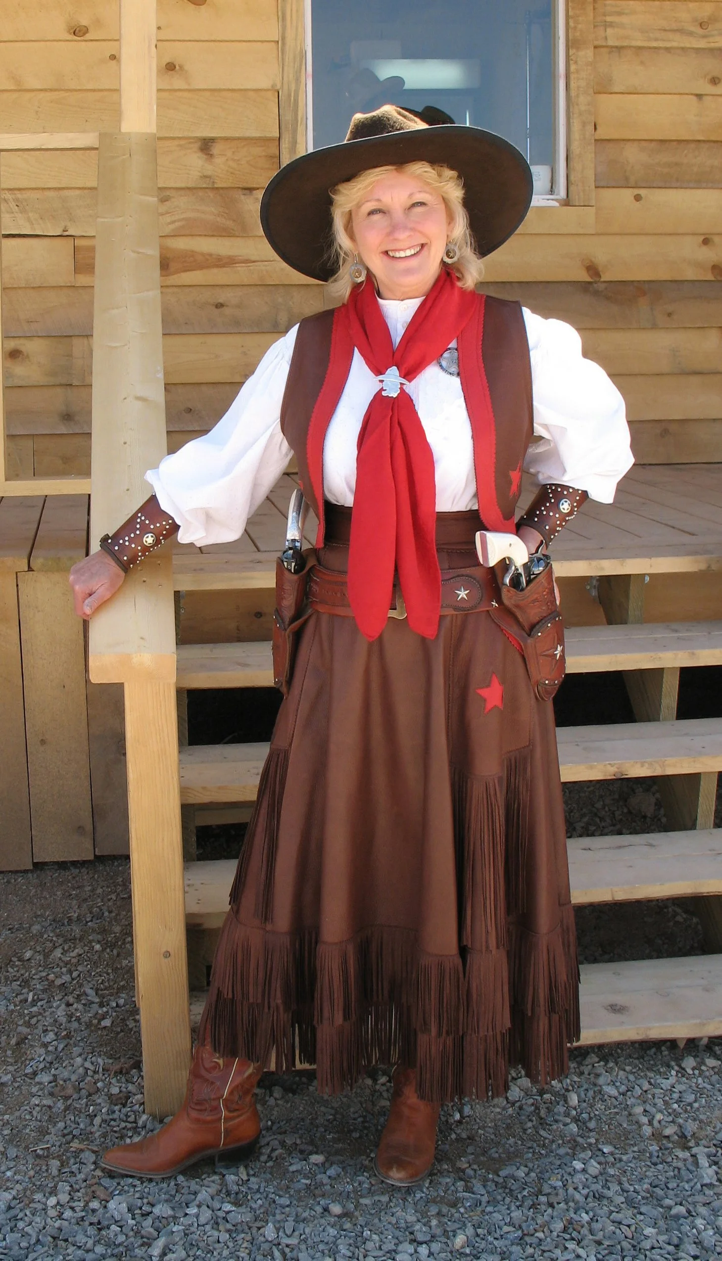 An Old West cowgirl wearing a wide-brimmed hat, white shirt, leather vest and skirt, red neckerchief, and boots, standing in front of wooden stairs and building.