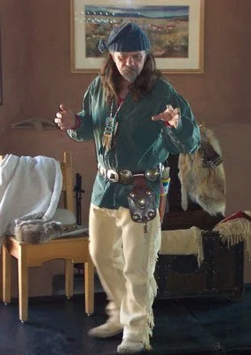 Mike Guli, in a Mountain Man outfit of toque, green cloth shirt, buckskin pants, moccasins,  beaded accessories and concho belt and pouch, tells stories of the Fur Trade Era at a program.