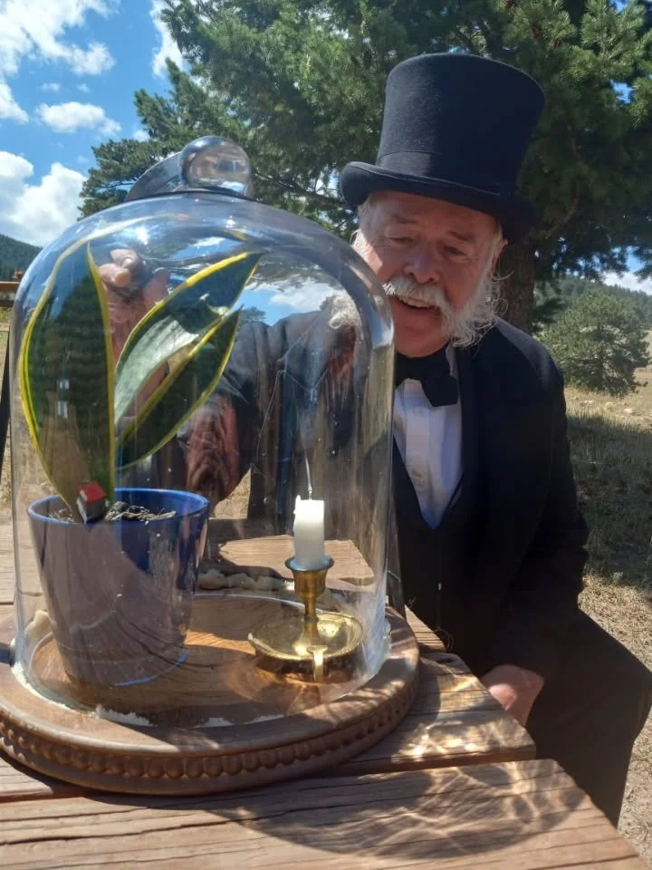 A man dressed in a vintage black suit and top hat, smiling and looking at a glass dome covering a potted plant with yellow-edged green leaves and a miniature candle on a brass holder, all on a wooden table outdoors under a blue sky with some clouds and trees in the background.