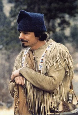 Mountain Man Mike Guli in buckskin outfit with beading, wearing toque, leaning on flintlock rifle.