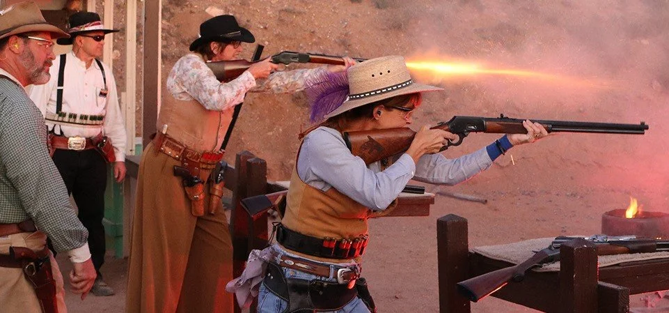 Women shooting rifles at a firing range, with a man observing nearby, in a desert setting.
