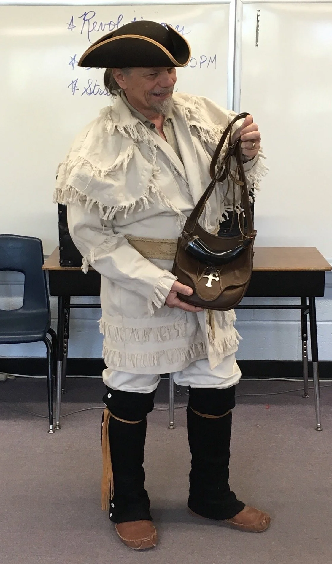 Mike Guli dressed in colonial-era costume, with a tricorn hat and fringed coat standing in a classroom setting, holding a leather bag with a metal cross ornament.