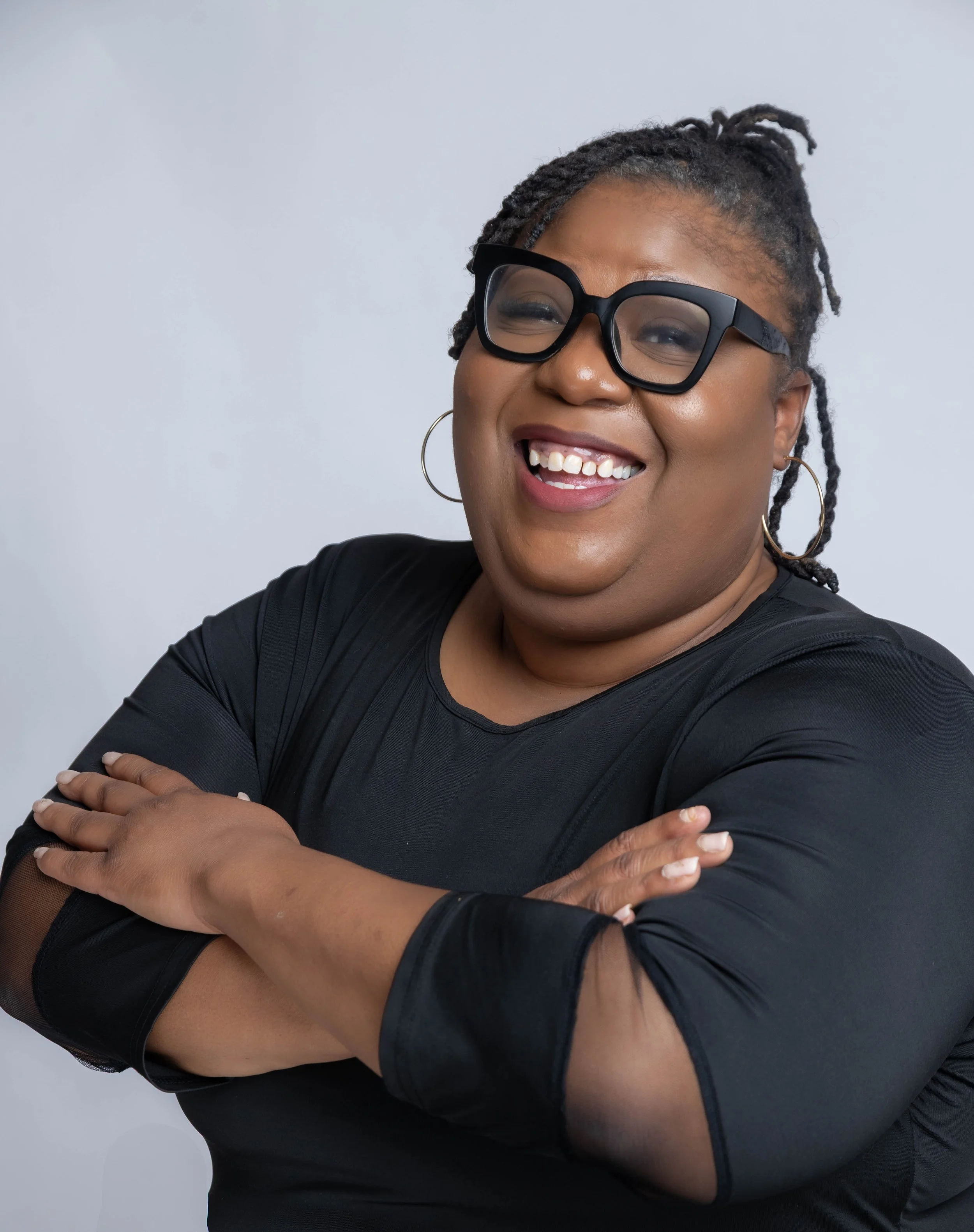 A smiling woman with glasses, hoop earrings, wearing a black top, arms crossed, against a plain light gray background.