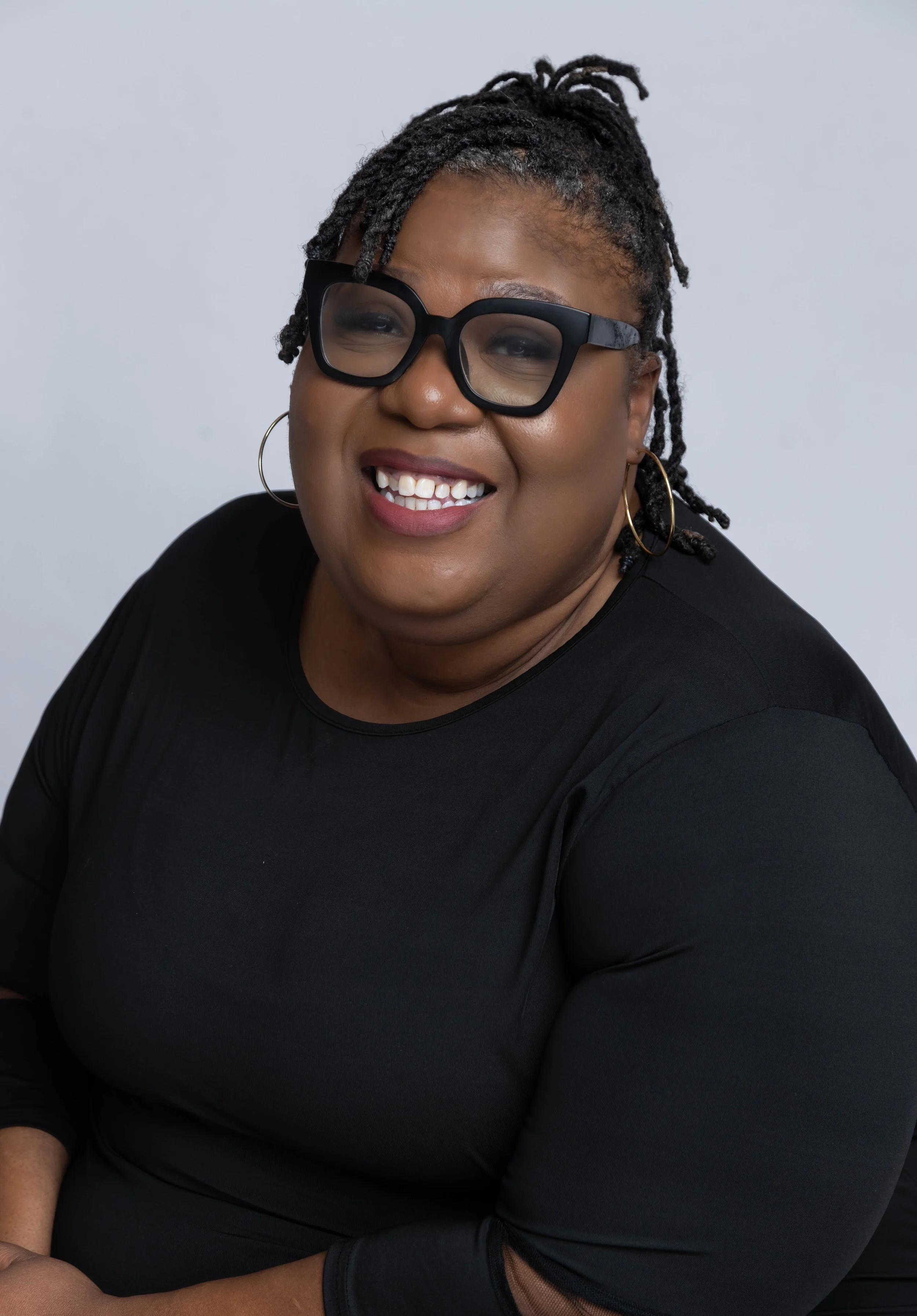 Portrait of a smiling woman with glasses, hoop earrings, wearing a black top, against a plain background.