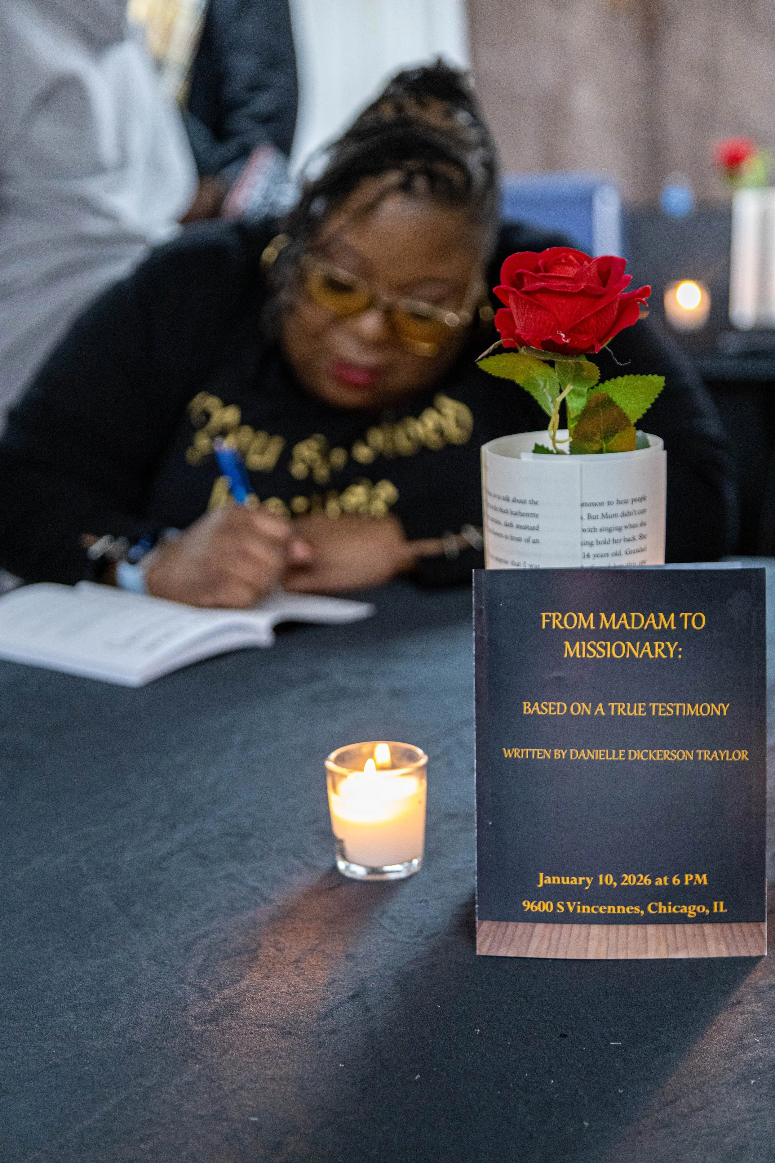 A woman seated at a table during a memorial event, signing a book with a red artificial rose in a white vase and a lit candle on the table. A sign on the table provides event details.