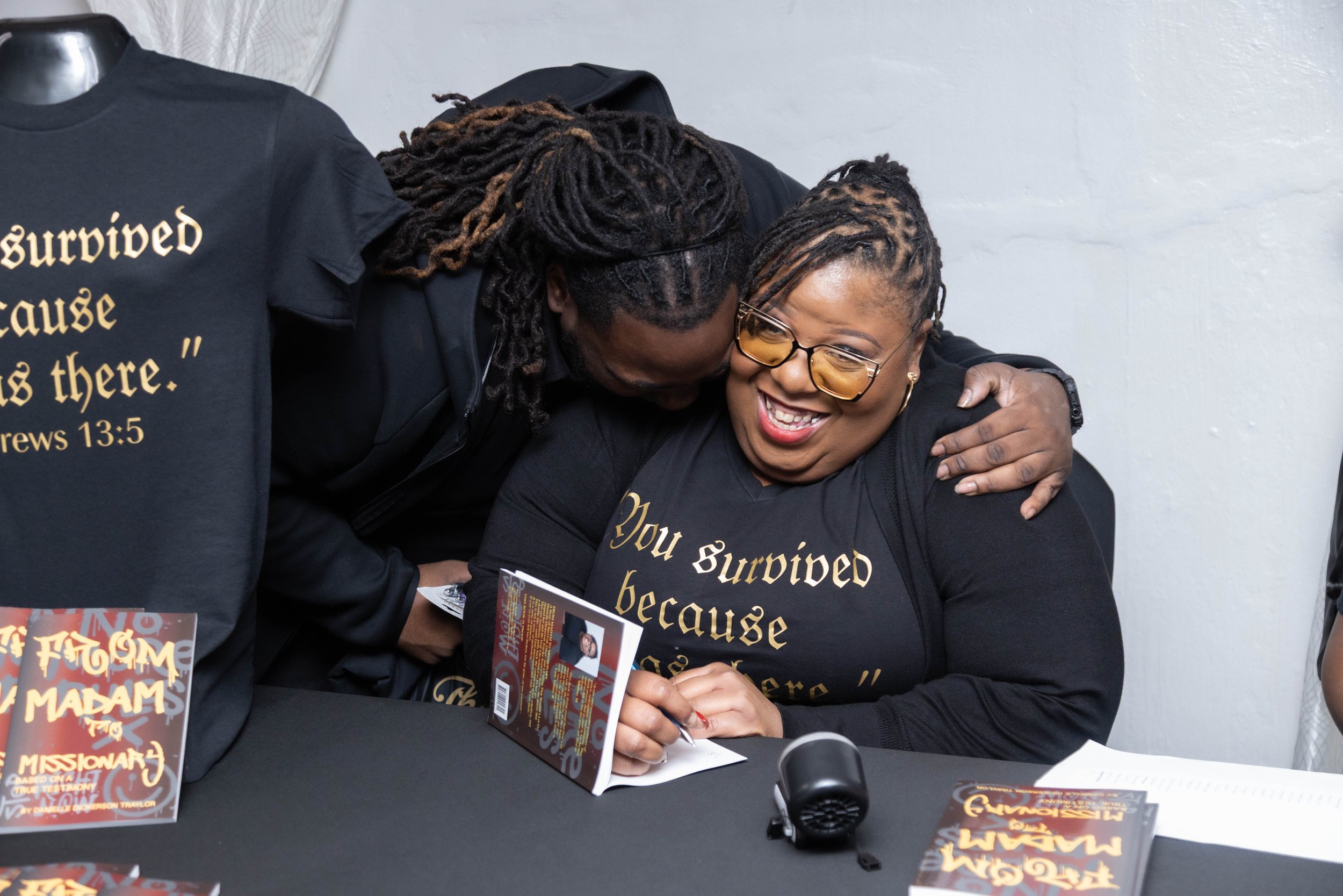 A woman with glasses is smiling while signing a book, with a man hugging her and whispering in her ear. They are at a table with copies of a book titled 'Fighting Madame Missionary.' The woman is wearing a black shirt with gold text that reads 'You survived because...'.