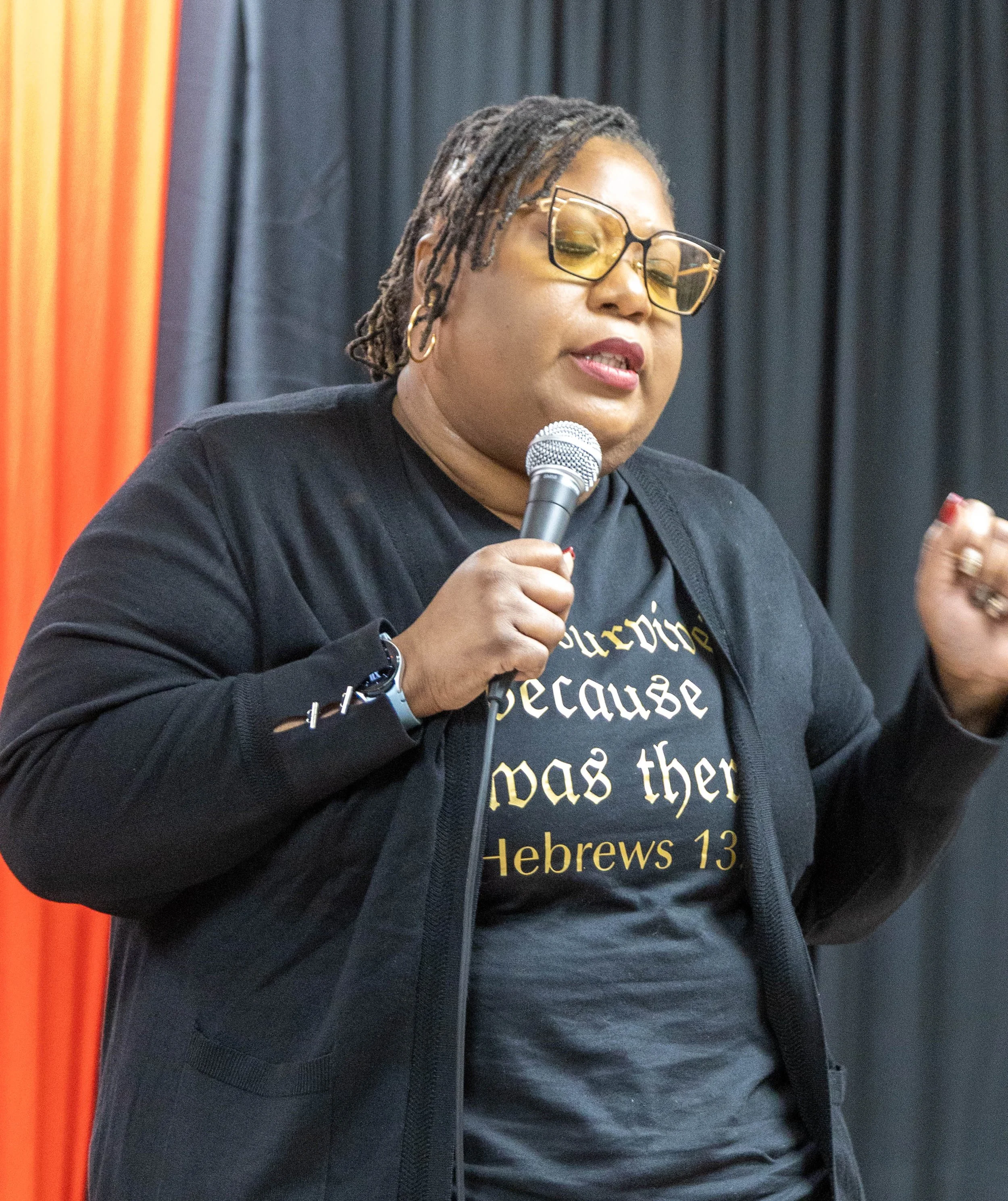A woman with glasses and dreadlocks speaking into a microphone at an event, wearing a black T-shirt with gold text that reads 'Hebrews 13' and additional text.