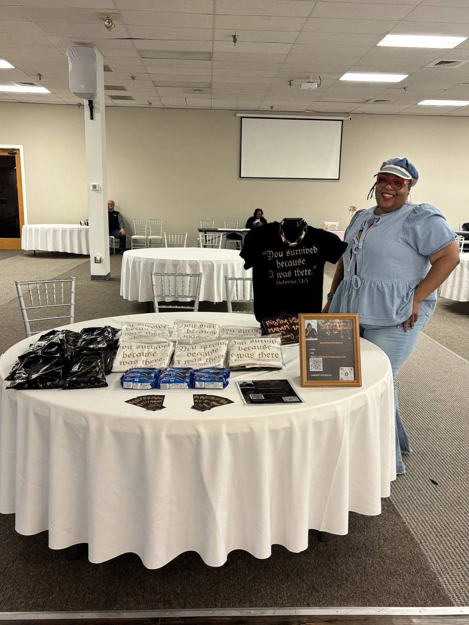 A woman standing next to a display table with t-shirts, pamphlets, and merchandise at an event in a large room with round tables dressed in white tablecloths.
