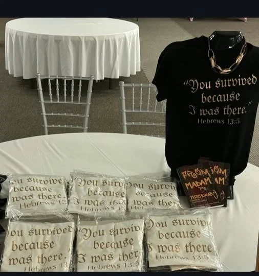 Display table with multiple clear bags containing messages, a black T-shirt with a religious quote, and a round table with a white cloth in the background.