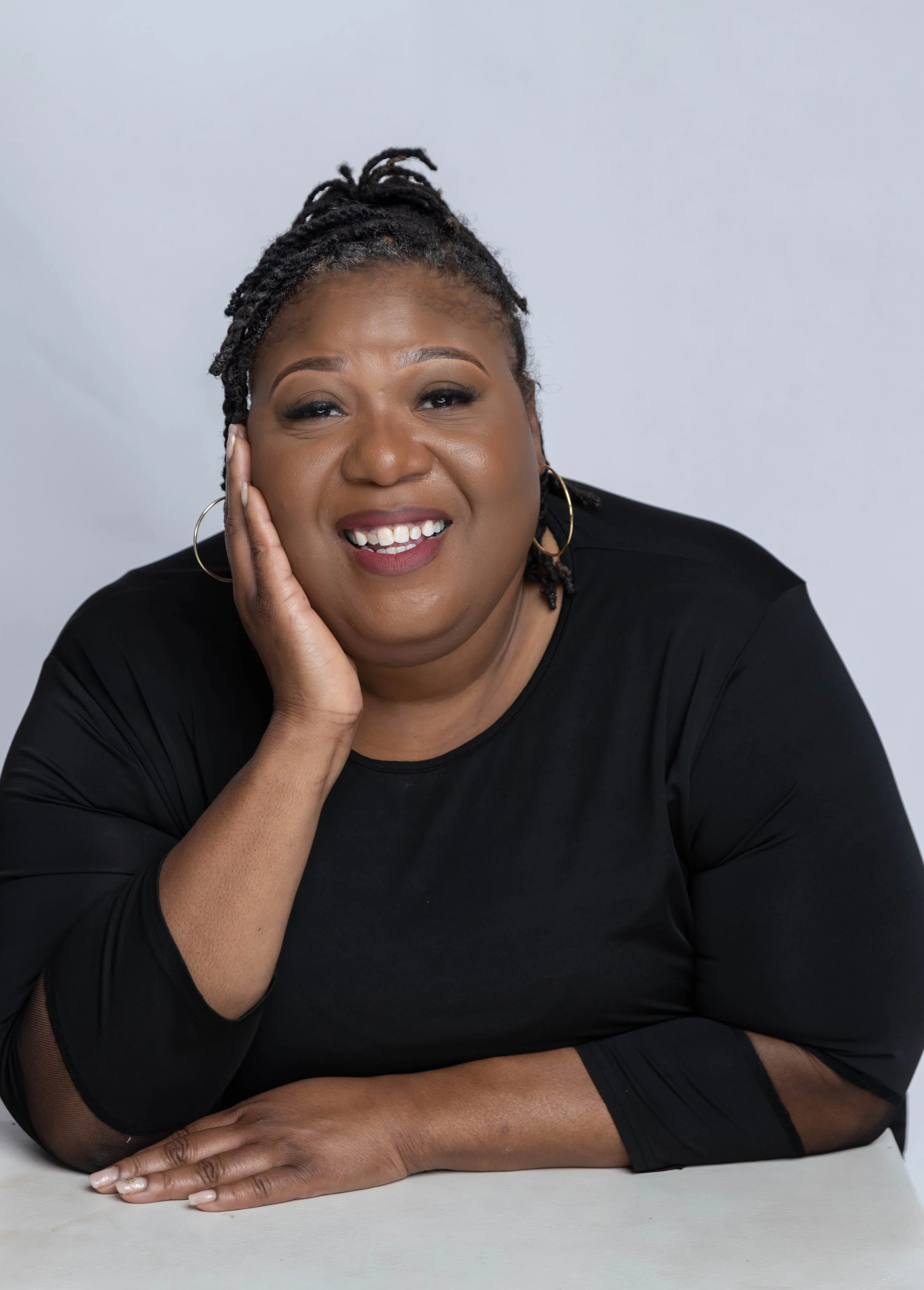 A smiling African American woman with dreadlocks, wearing a black top, sitting at a table with her chin resting on her hand, against a light gray background.