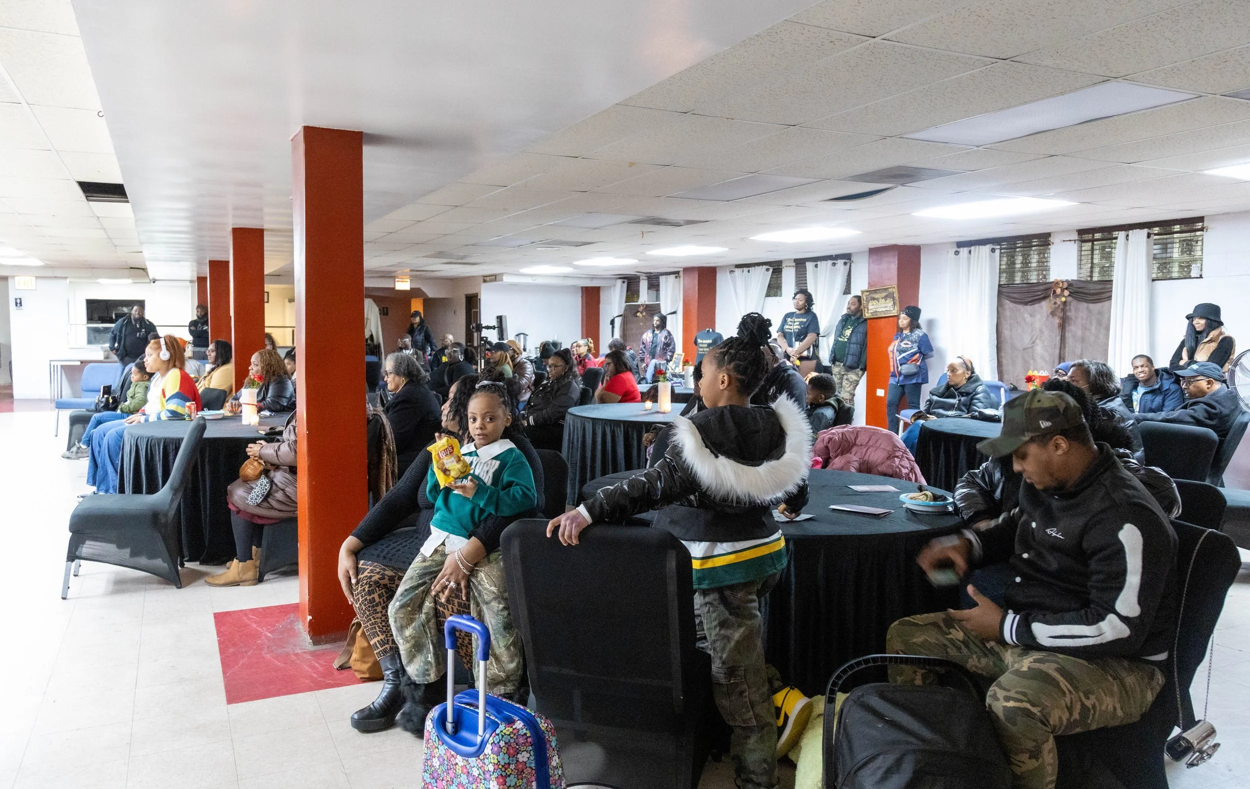 People seated at round tables in a large indoor event space, some standing against the walls, with a mix of adults and children, some eating, some using phones, and some looking towards the front of the room.