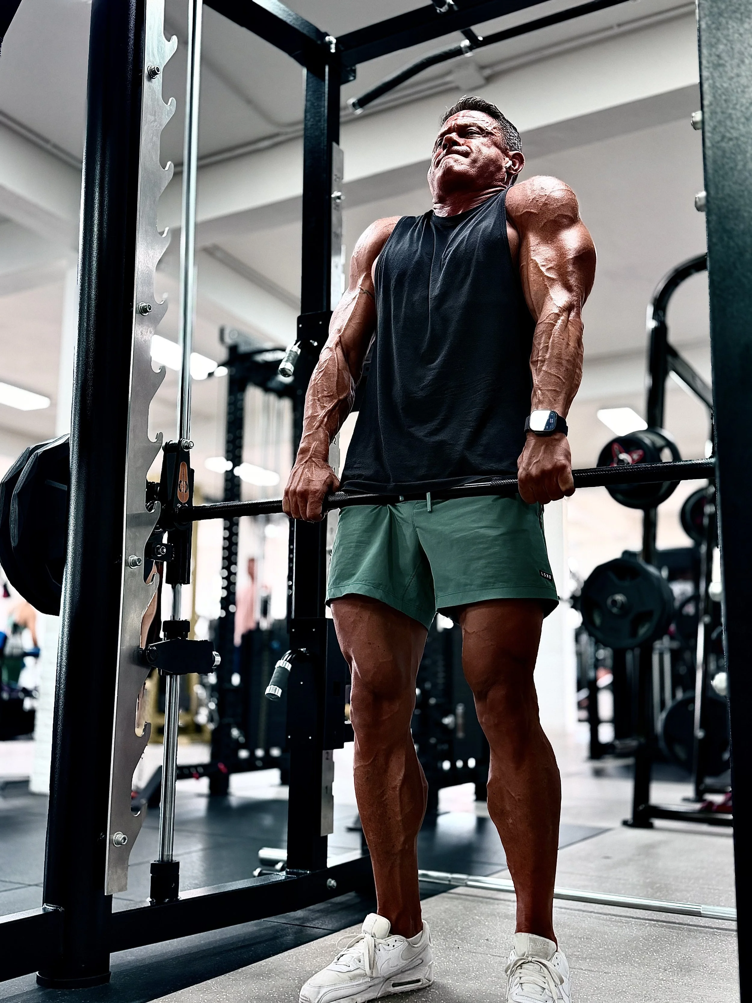 A muscular man lifting weights in a gym, wearing a black tank top, green shorts, white sneakers, and a smartwatch, with a focused and intense expression.
