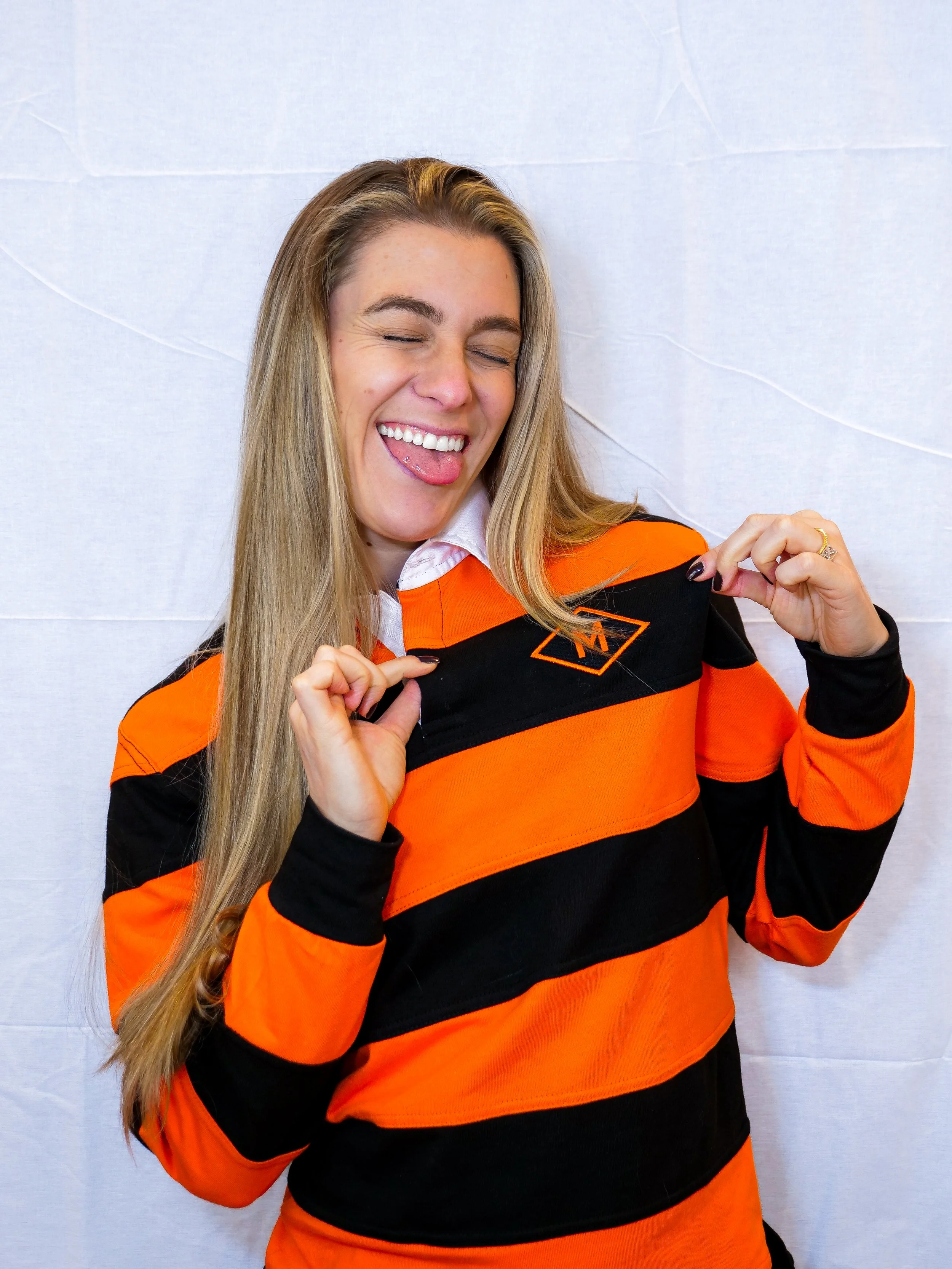 A woman with long blonde hair wearing an orange and black striped sports jersey, smiling and sticking out her tongue, holding the jersey's collar with both hands.