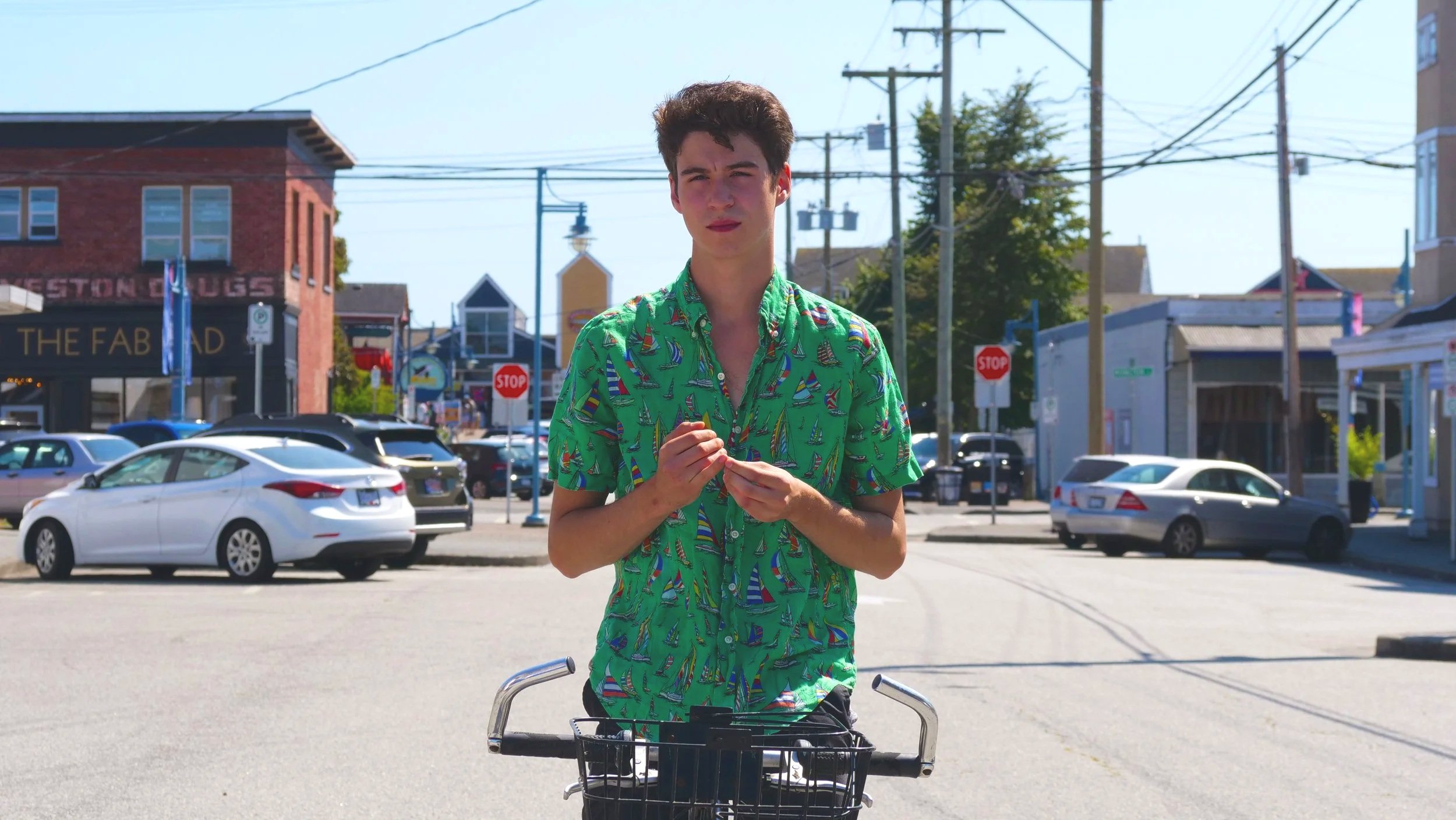 A young man with short dark hair wearing a green shirt with a sailboat pattern standing behind a bicycle on a city street with parked cars, buildings, and power lines visible in the background.