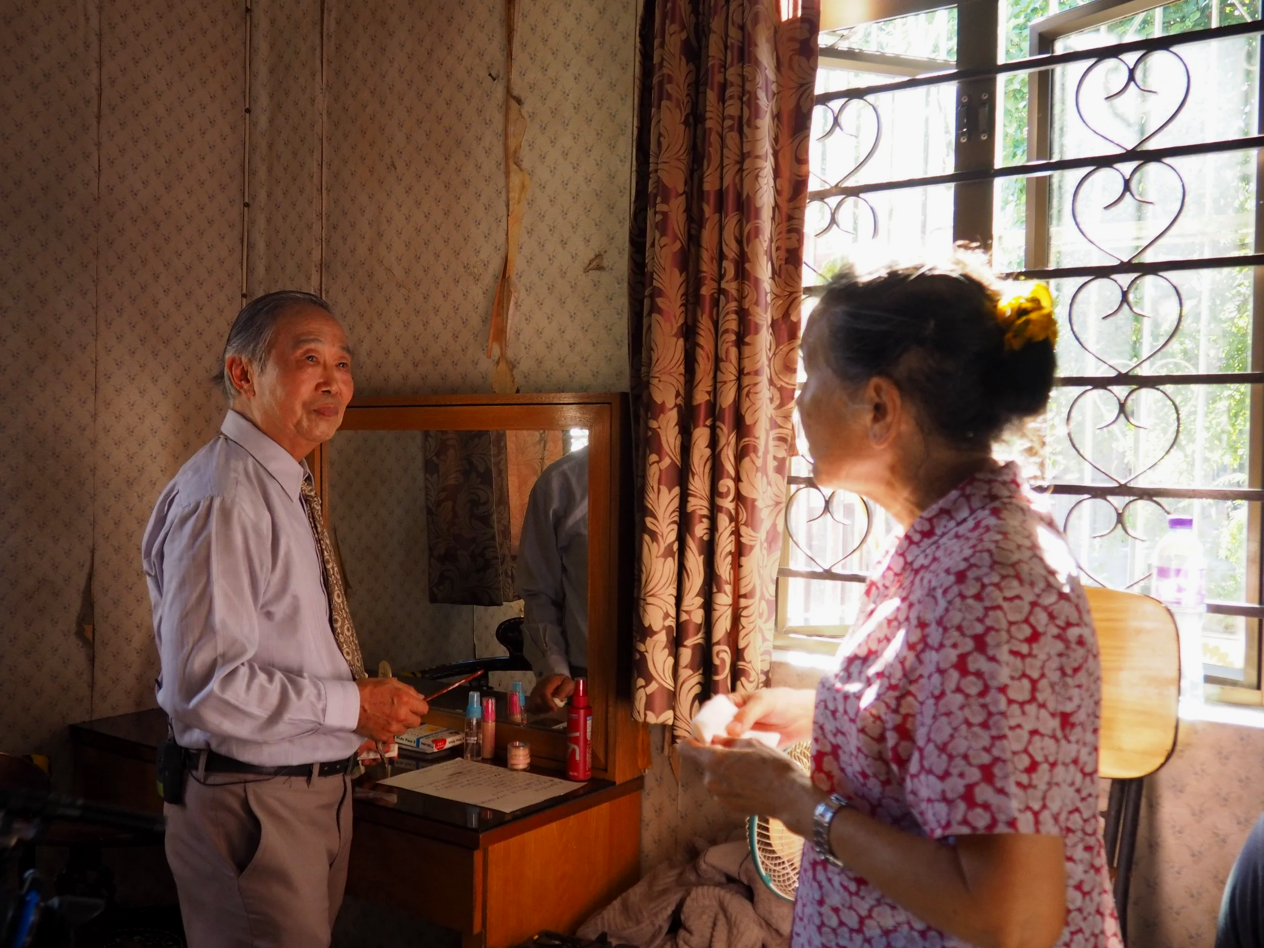 An elderly man and woman standing in a room near a window, the man is holding a makeup brush, and the woman is holding a tissue, with makeup items on a dresser.