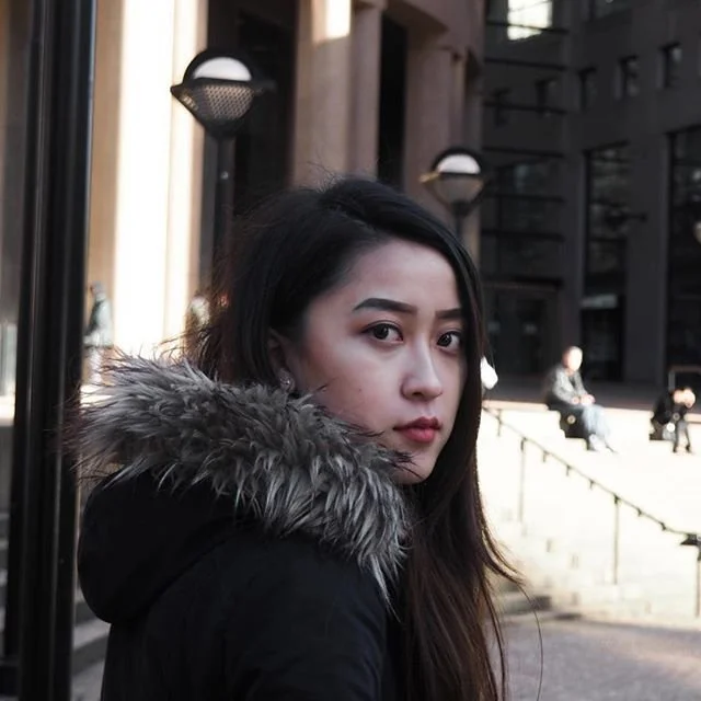 Young woman with dark hair and fur-lined coat looking over her shoulder outdoors in an urban setting.