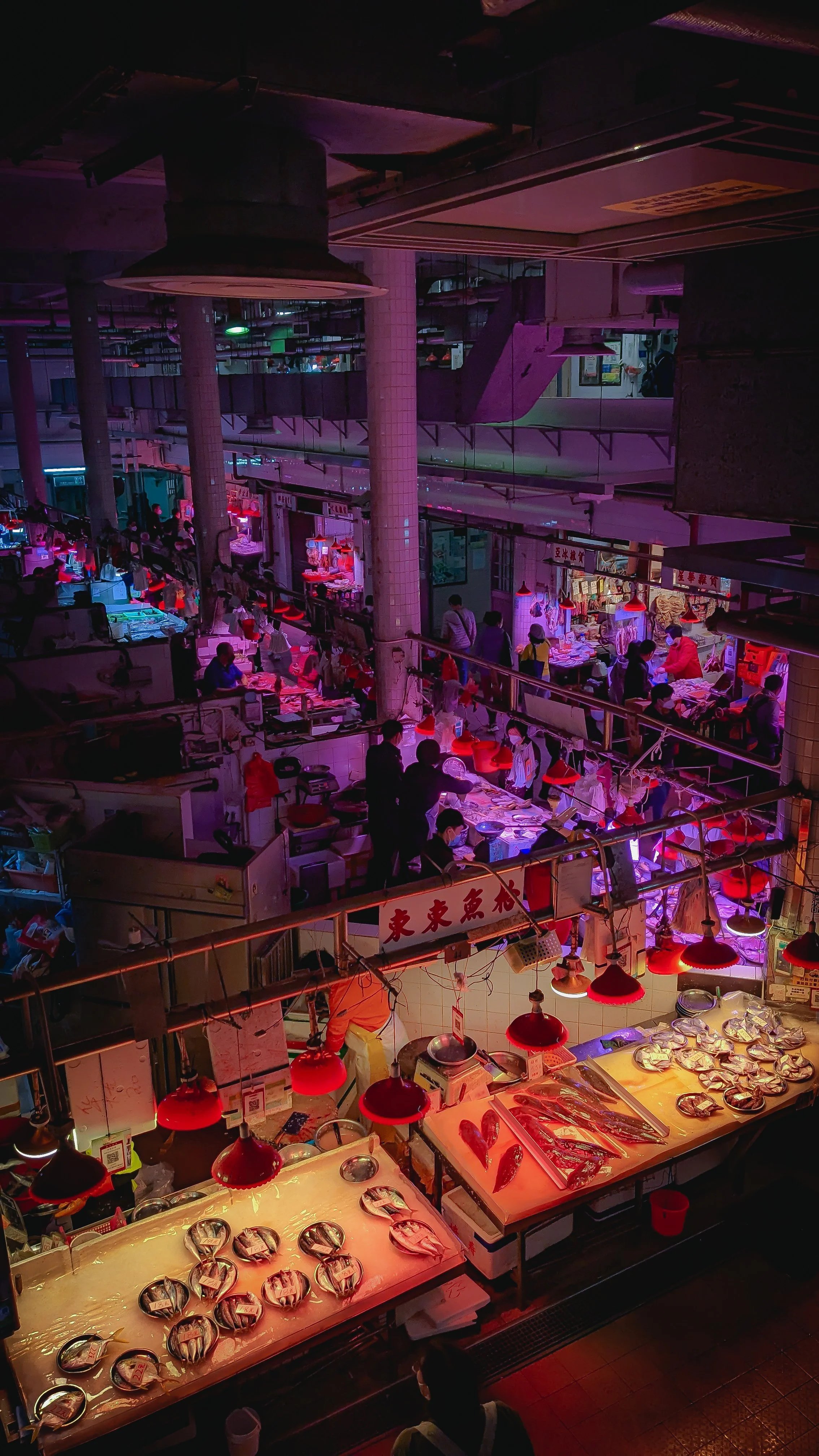 A bustling indoor market or food court with various food stalls and vendors selling seafood, illuminated with colorful and red lighting.