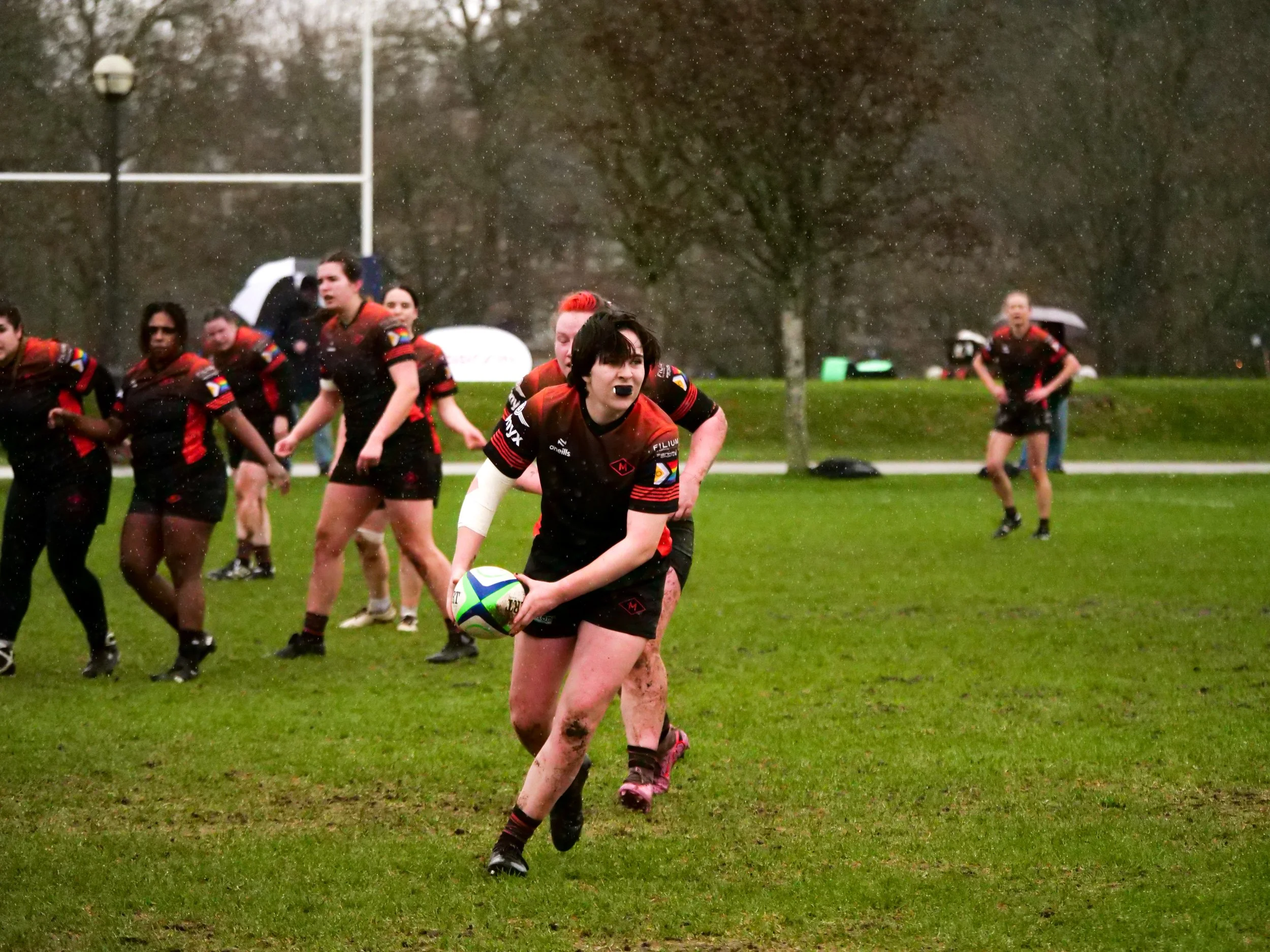 A women's rugby team playing in a muddy field on a rainy day. One player in the foreground is holding a rugby ball, running, and looking determined. Other players are behind her, some running, some standing. The background shows trees, goalposts, and
