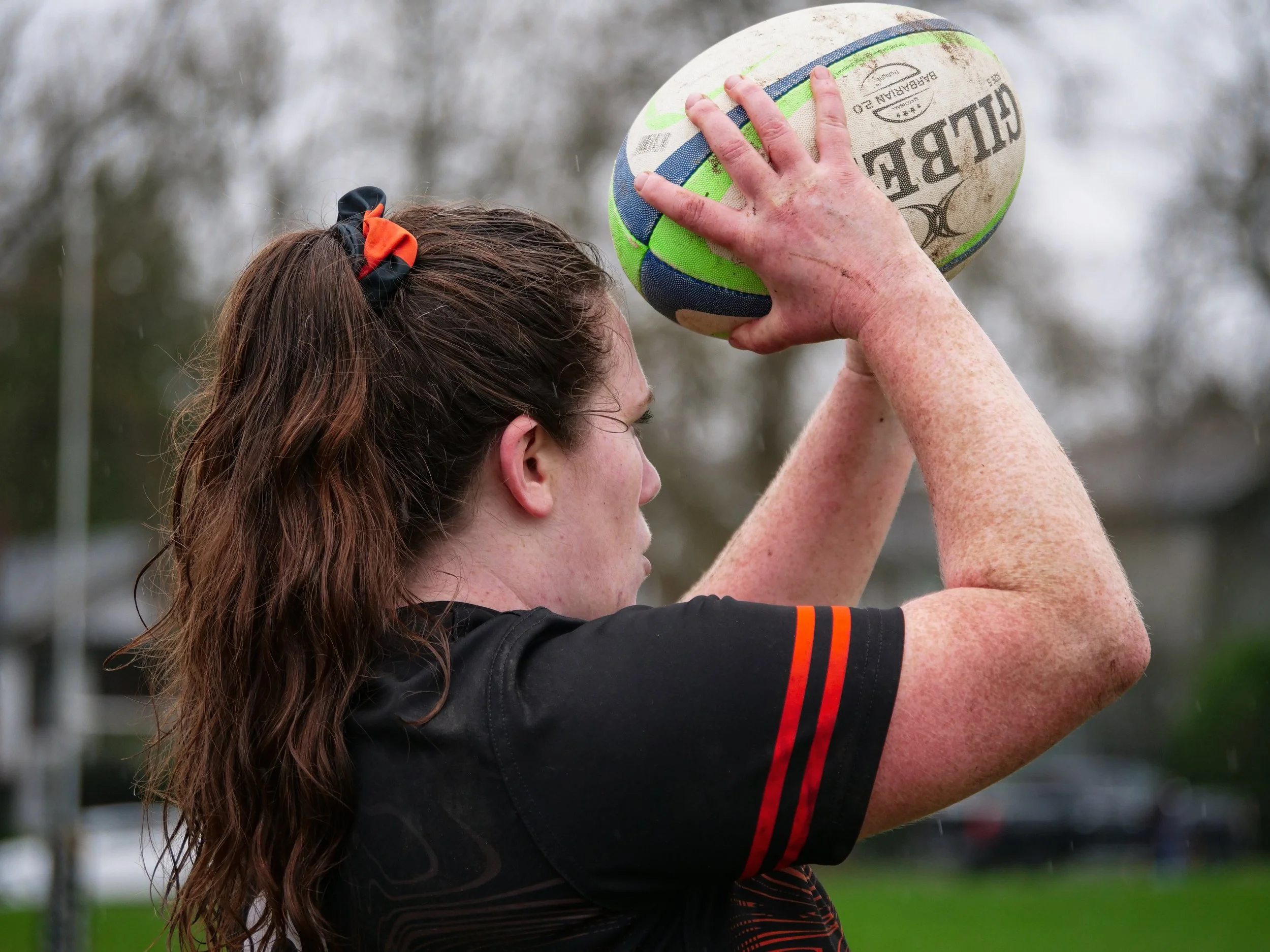 A woman with long brown hair tied with a black and orange scrunchie preparing to throw a rugby ball in a park.