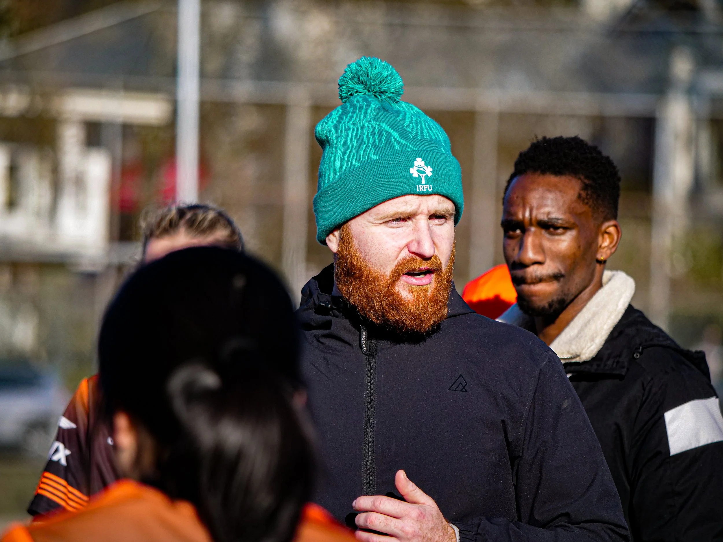 Man with red beard wearing a teal beanie and black jacket speaking to a group outdoors.