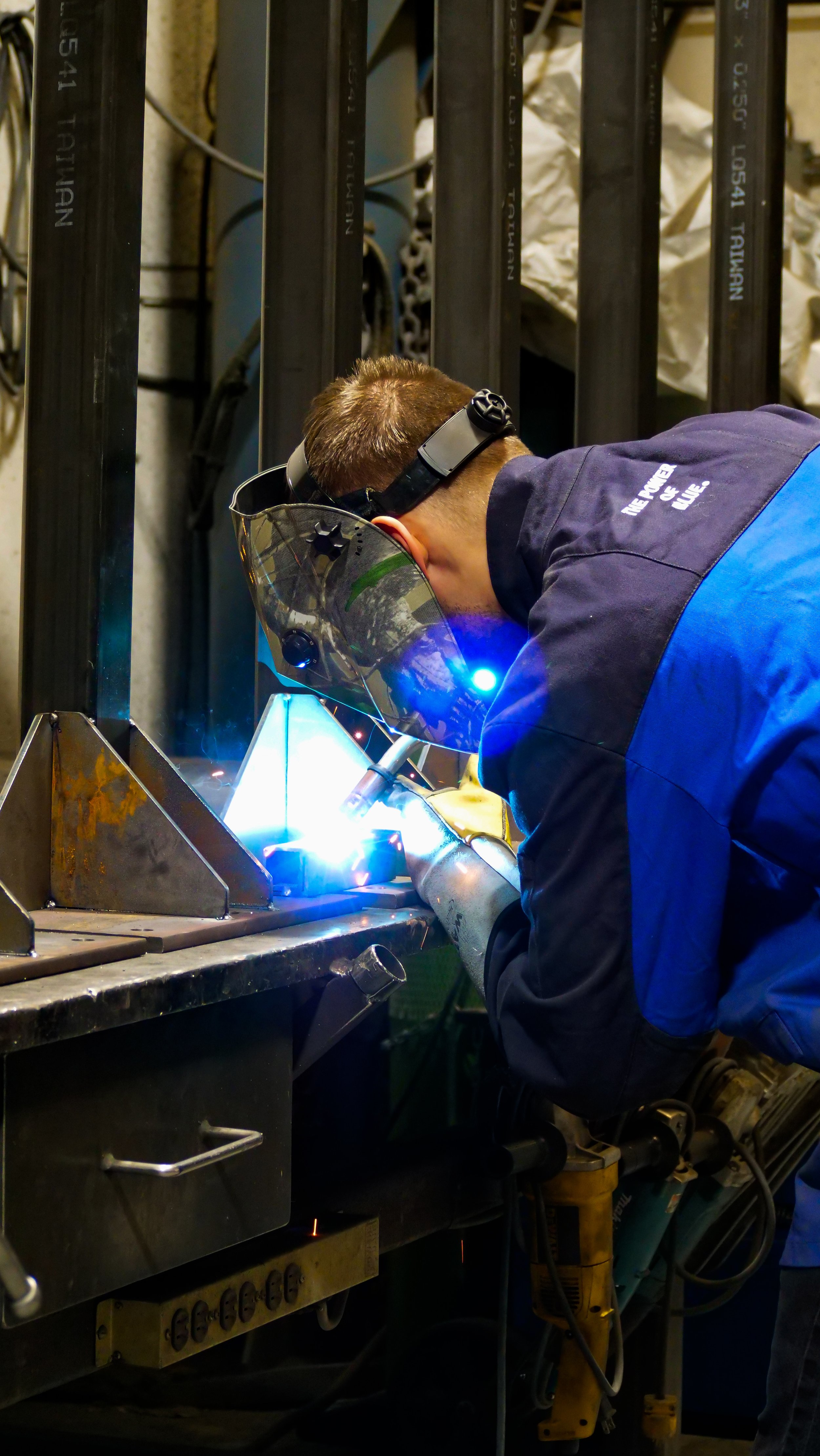 A person welding metal components in an industrial workshop. They are wearing a protective welding helmet, gloves, and a blue and black work uniform.