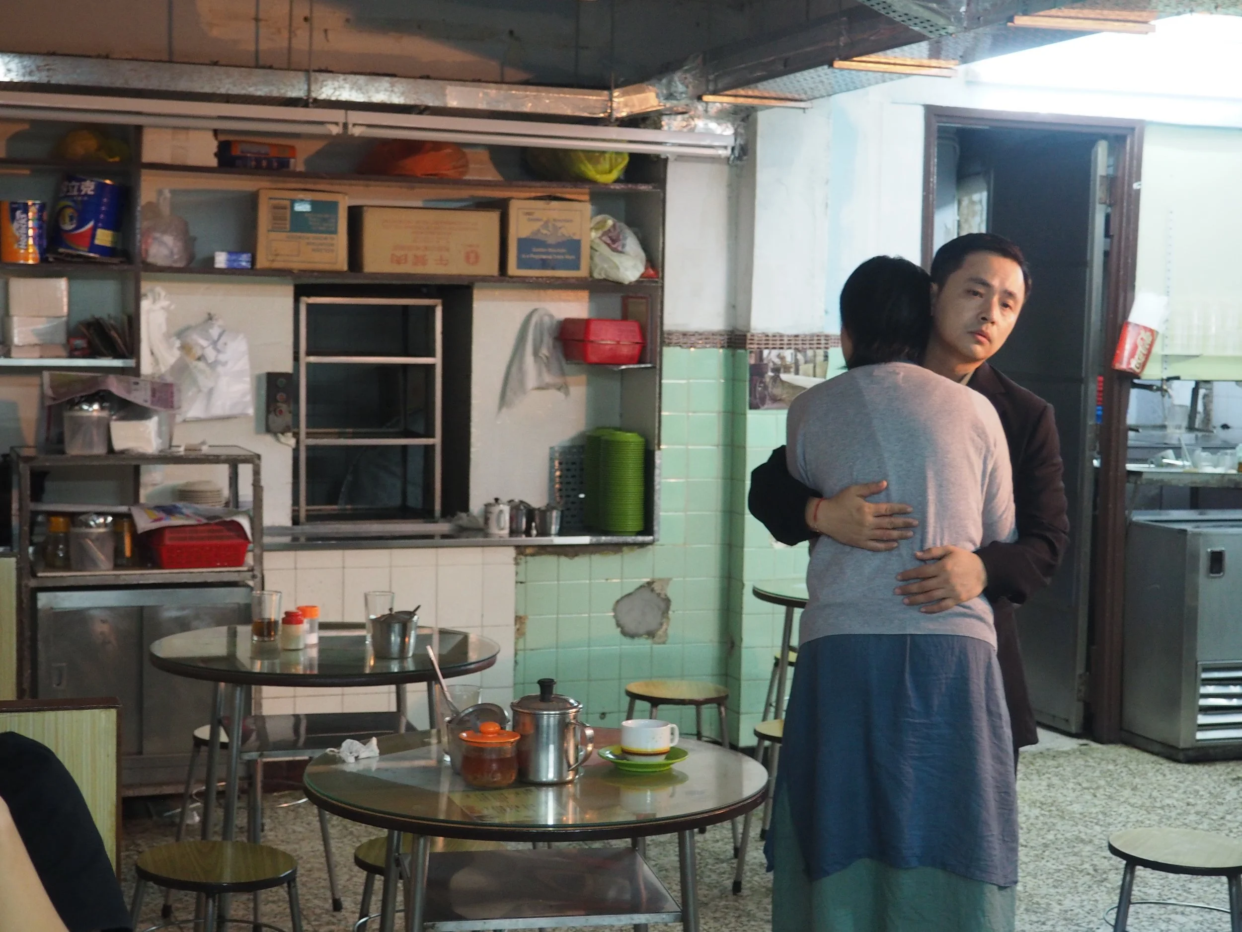 A man and a woman embrace in a small, cluttered restaurant or cafe with green tiled walls, tables, and kitchen equipment in the background.