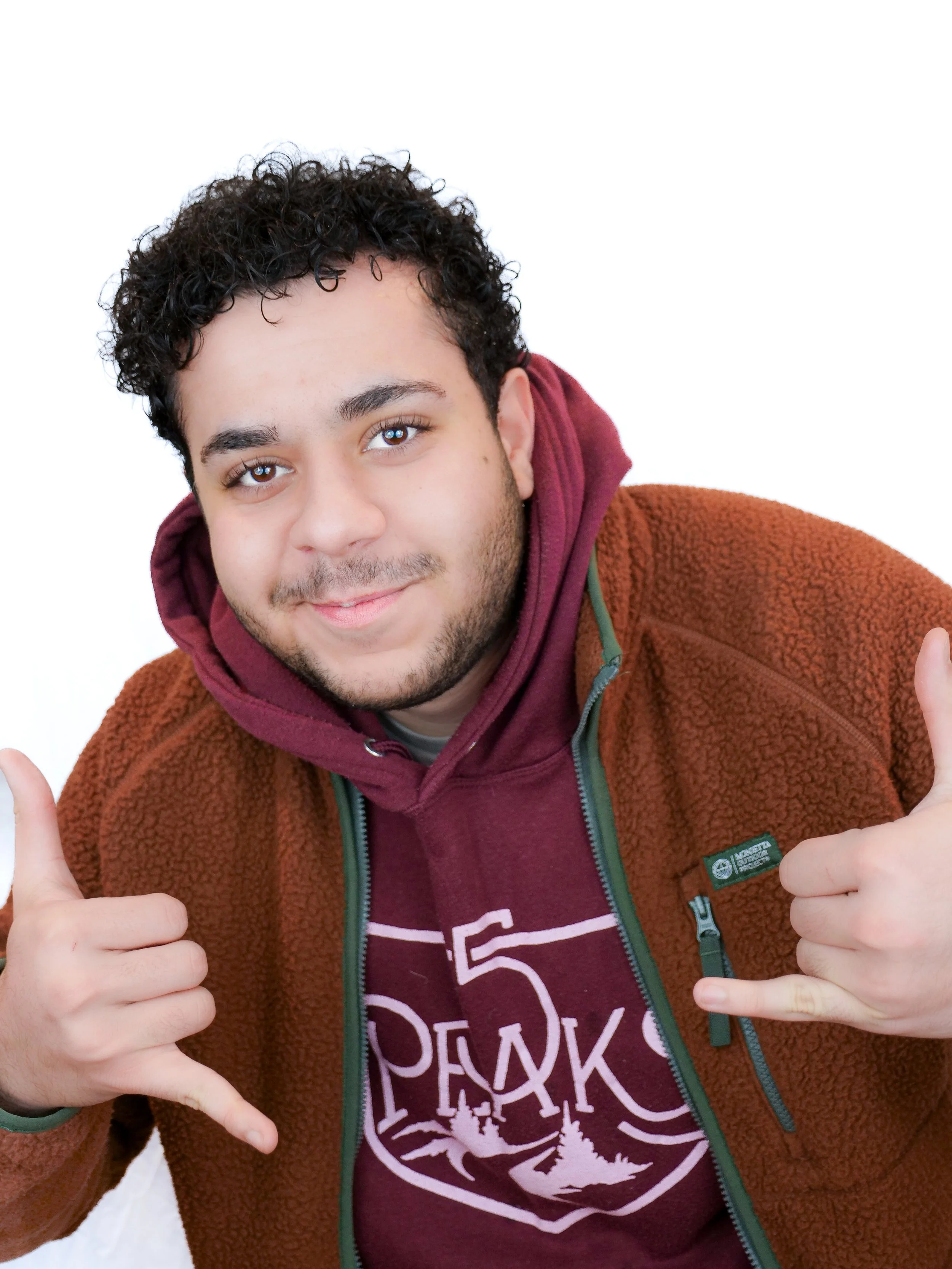 Young man with curly dark hair and light skin giving thumbs up and shaka sign, wearing a maroon hoodie and a brown fleece jacket, smiling against a white background.