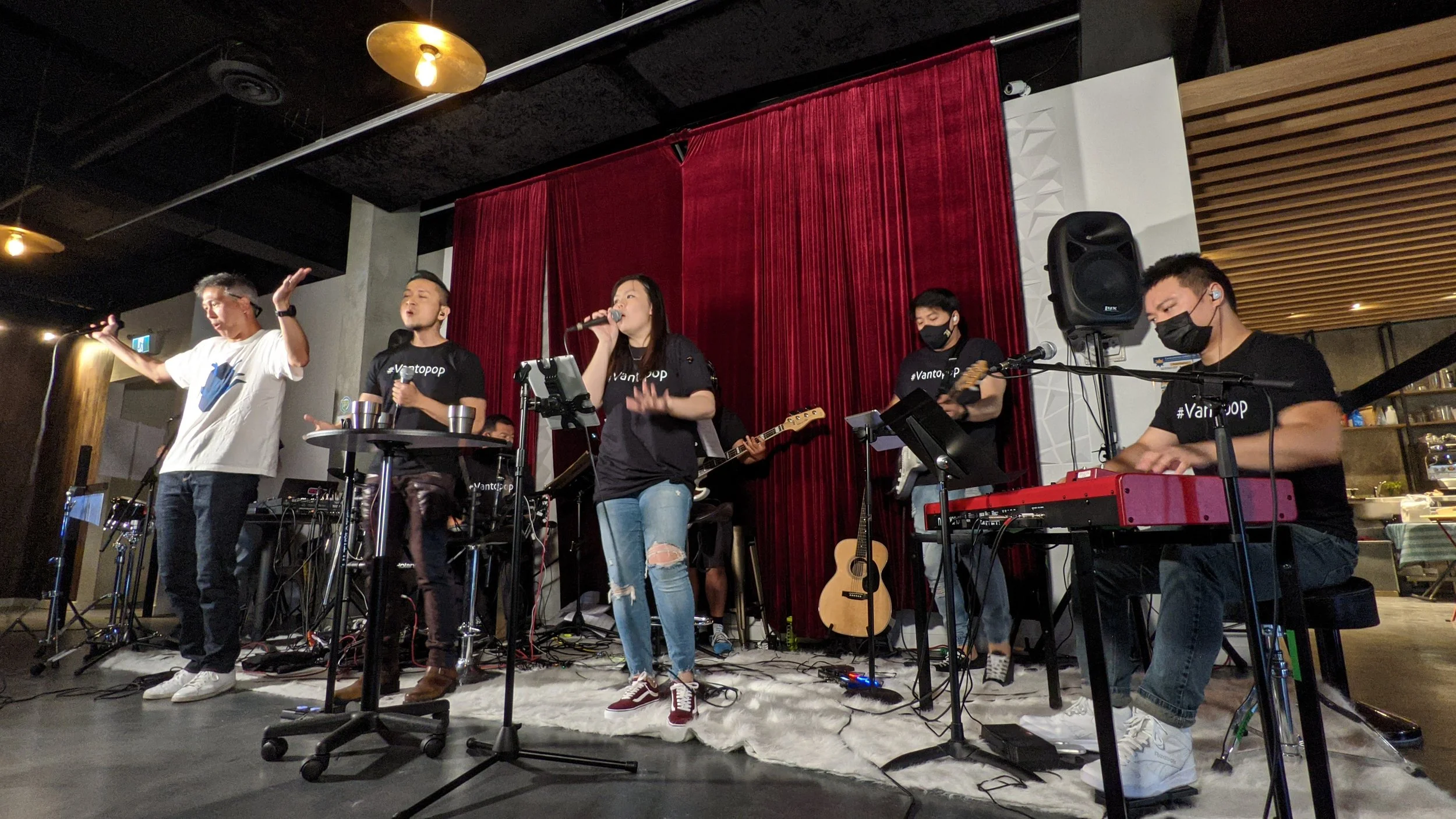 A band performing on stage with five members, some wearing black shirts with the hashtag '#Vantopop', in a room with red curtains, gold ceiling lights, and wooden accents. One person is singing into a microphone, another playing a keyboard, and other