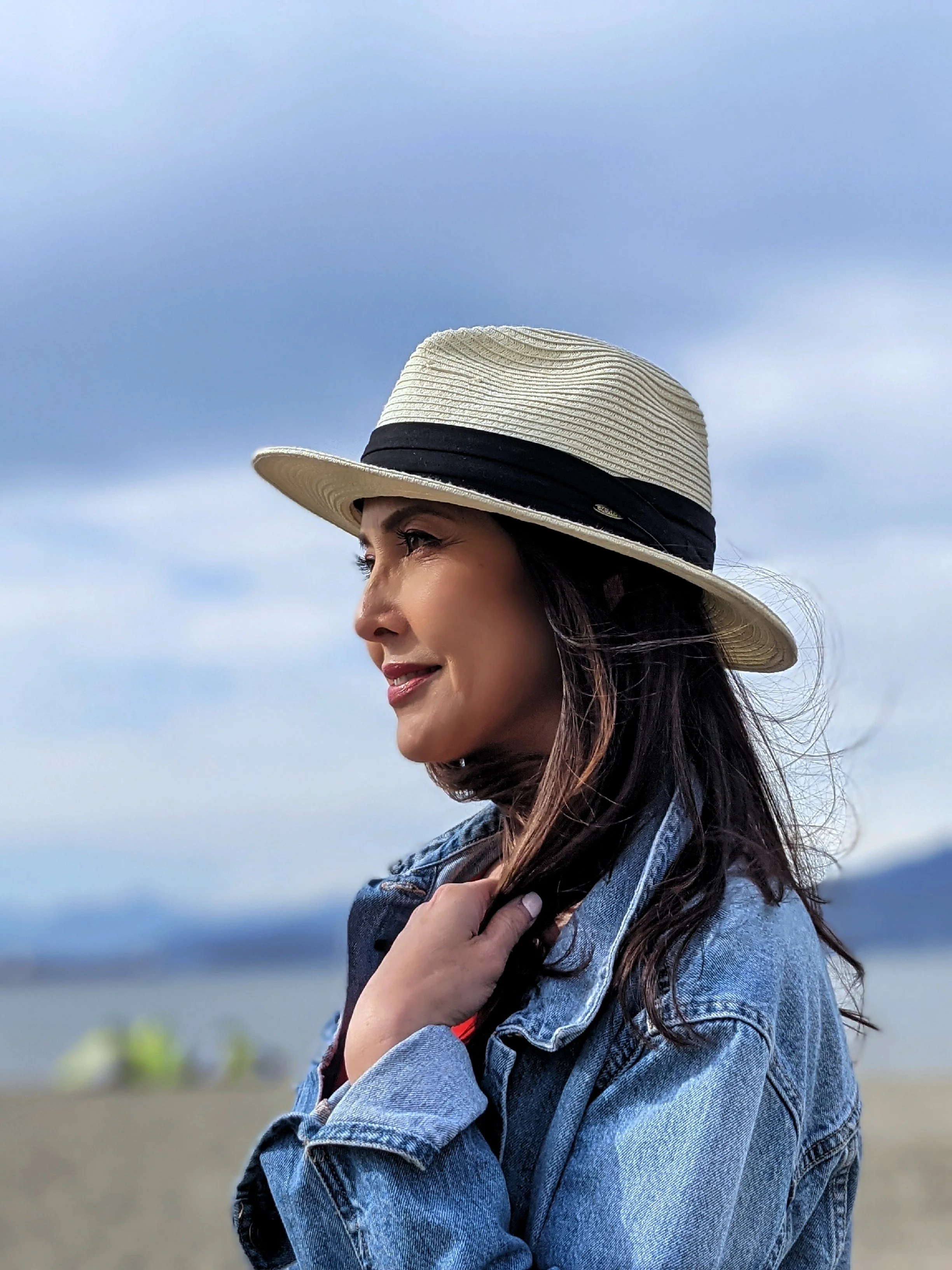 Woman wearing a straw hat with black band and denim jacket standing outdoors with a cloudy sky and blurred natural landscape in the background.
