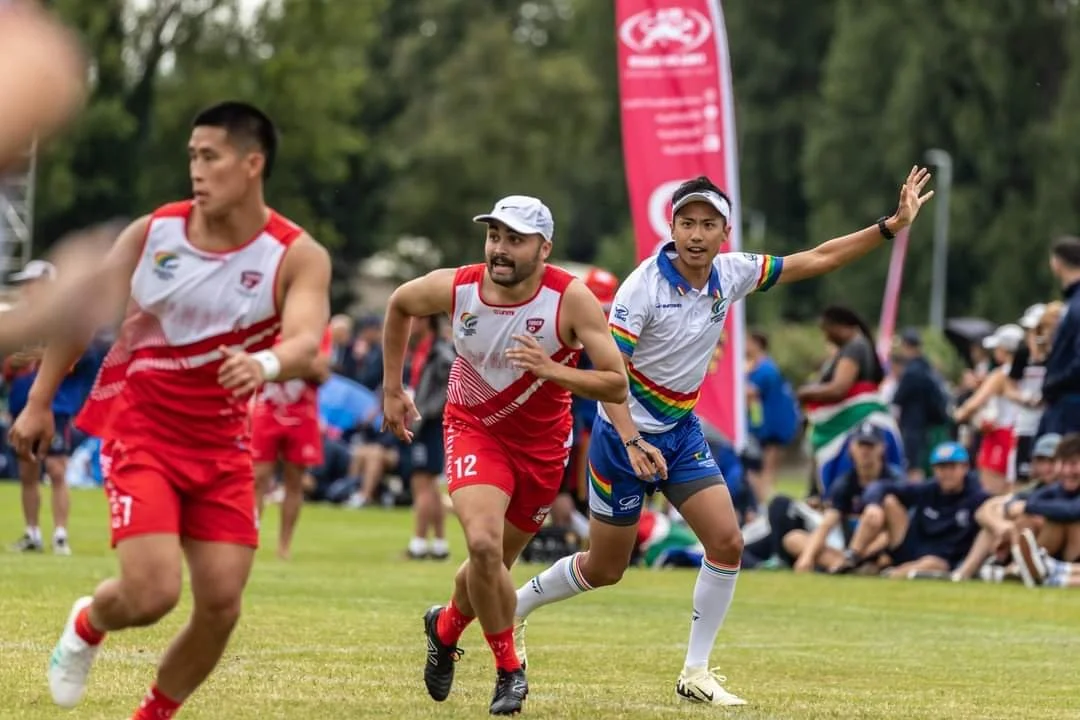 Three athletes running on a grass field during an outdoor sports event with spectators in the background.