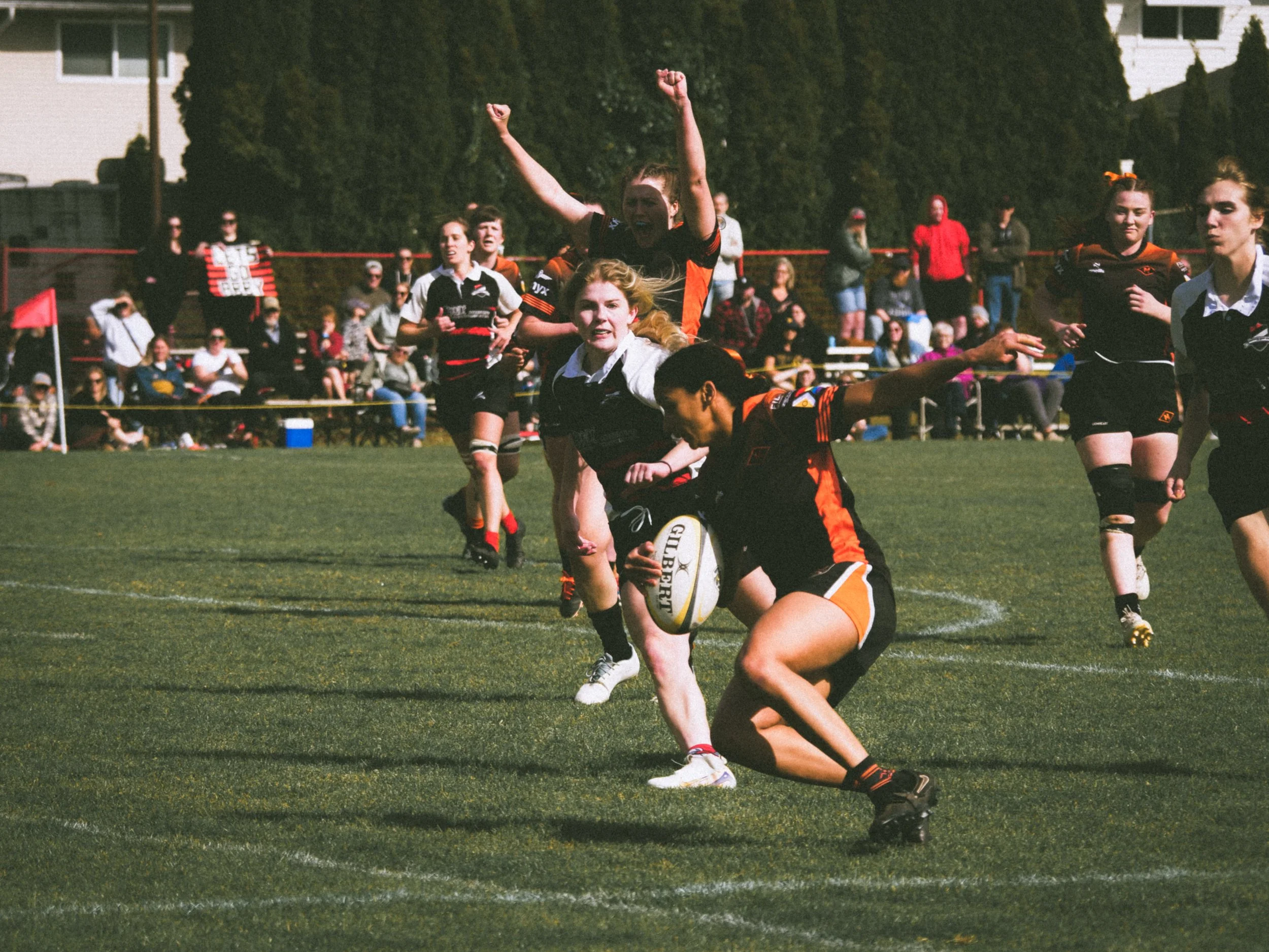 Women playing rugby on a grassy field during a game, with spectators watching in the background.