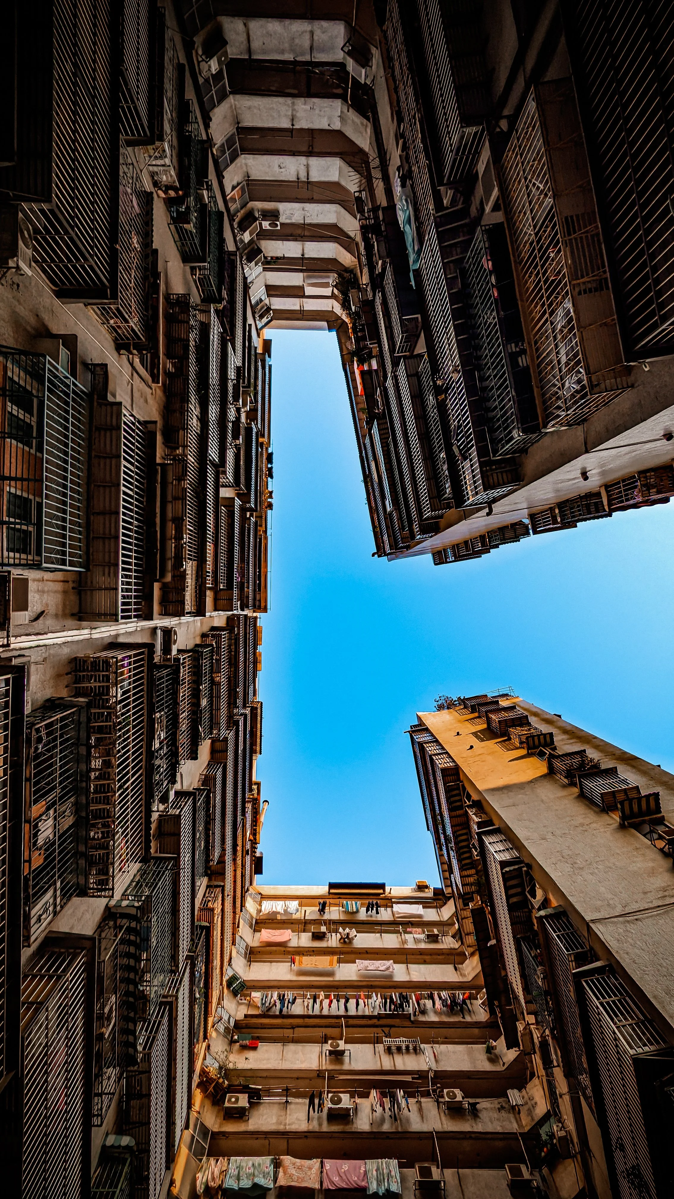 Looking up at a courtyard surrounded by tall apartment buildings with balconies, laundry hanging, and air conditioning units, with a clear blue sky overhead.