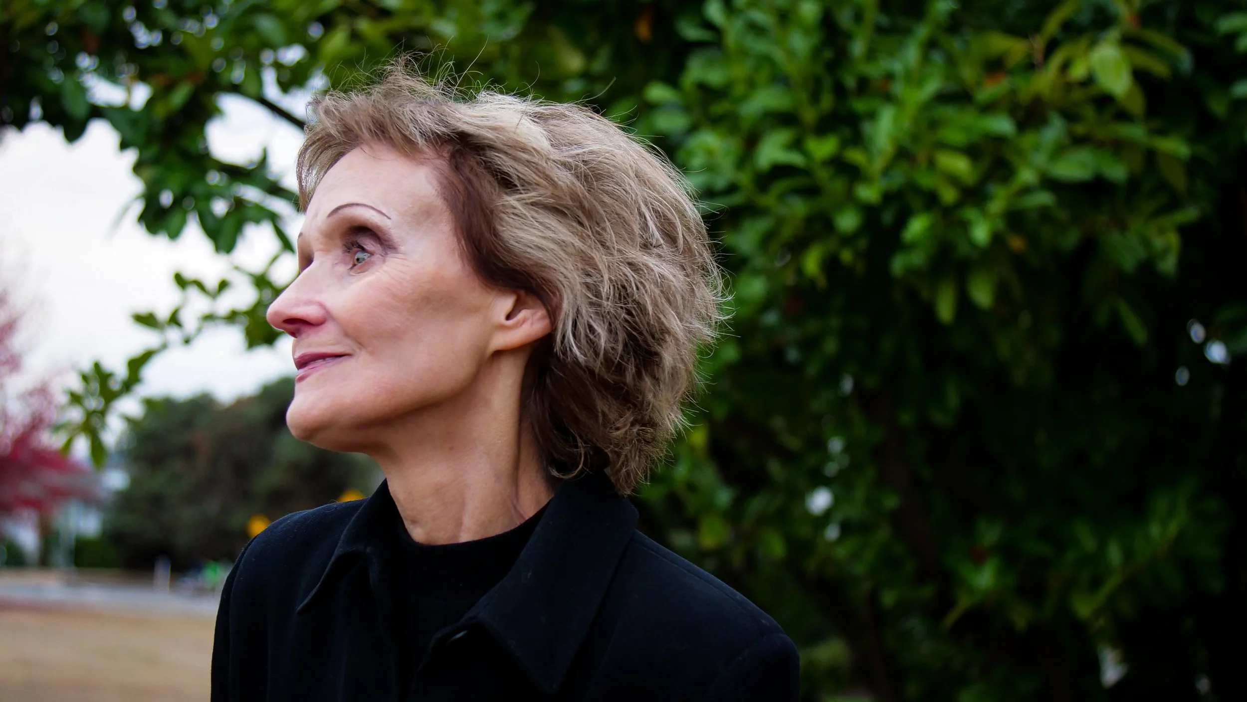 A side profile of an older woman with short, wavy, gray hair standing outdoors surrounded by green foliage.
