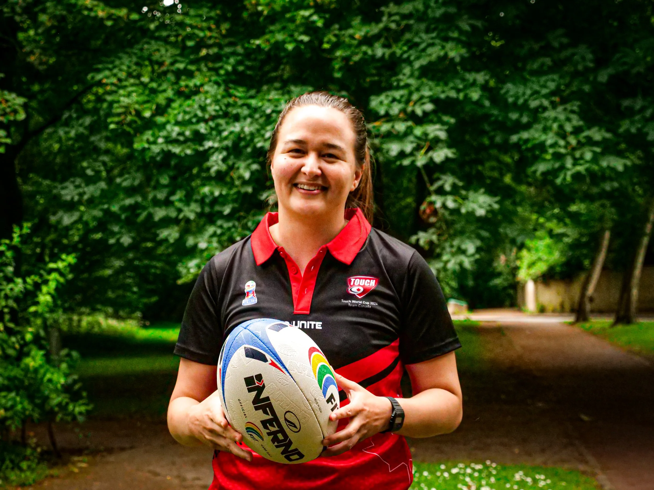 A smiling woman in a black and red sports shirt holding a rugby ball outdoors in a park with green trees in the background.