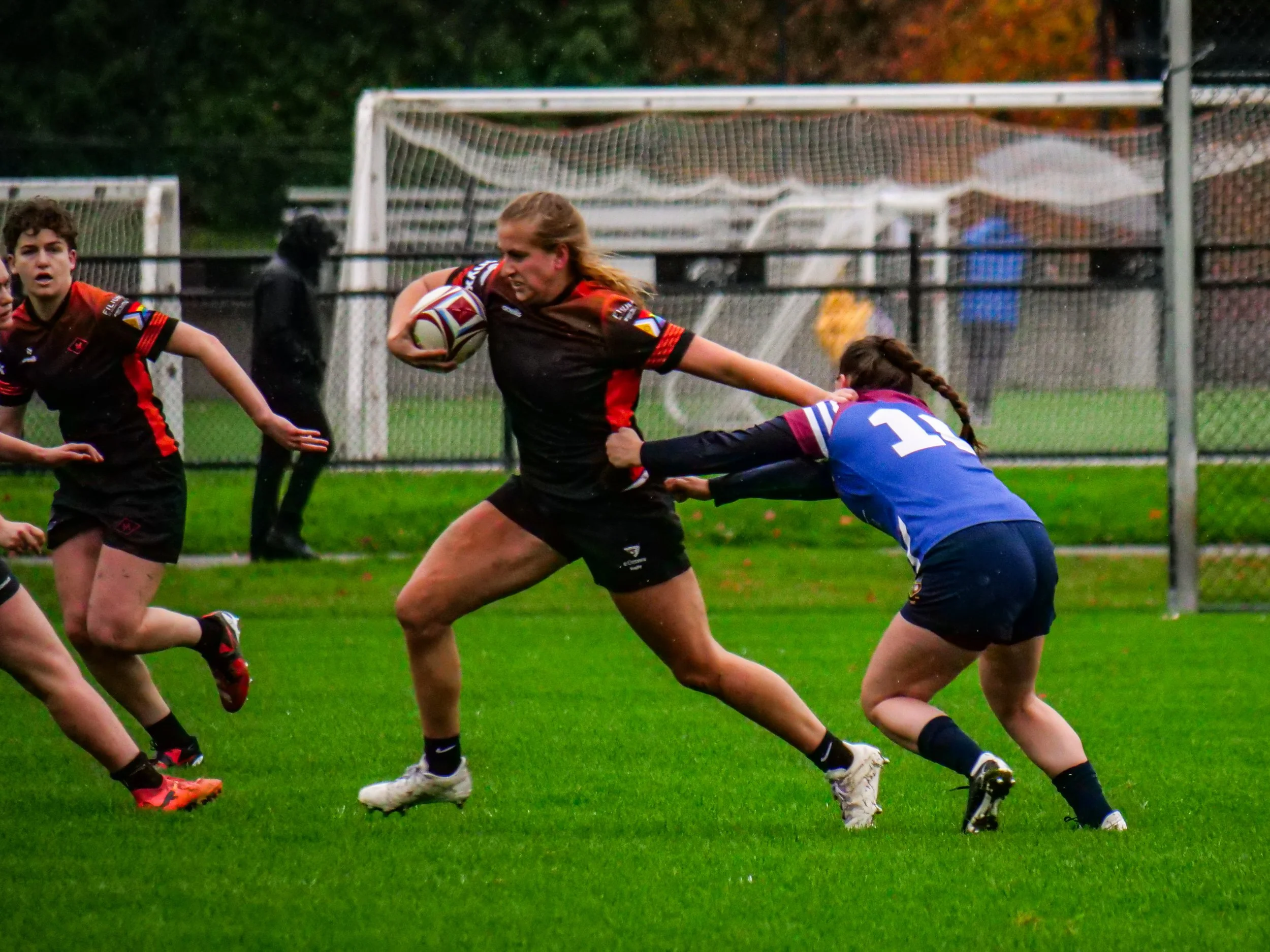 Rugby match with female players on a grass field; one player in a black and red uniform holding a rugby ball, running while being tackled by a player in blue and white uniform.