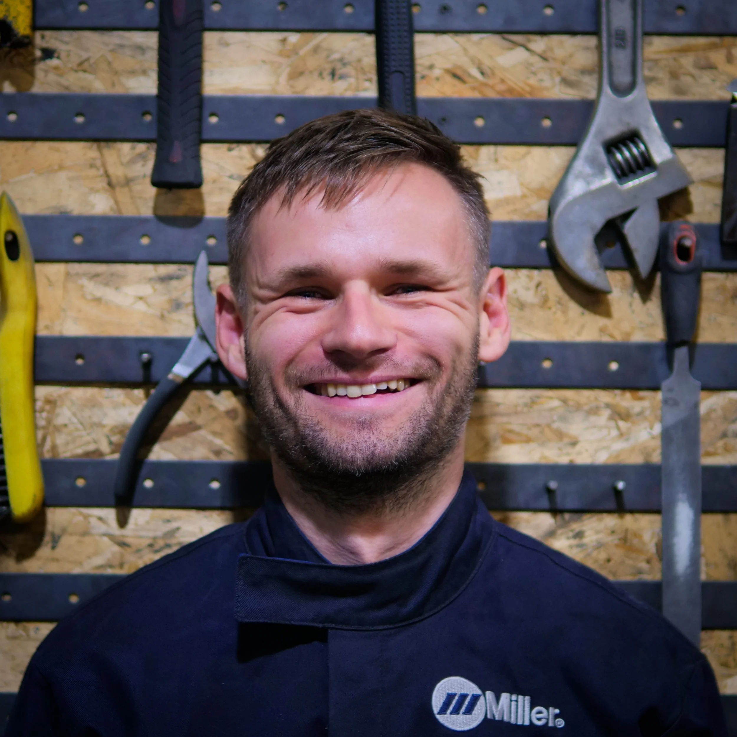 A smiling man with short brown hair and a beard, wearing a black Miller jacket, standing in front of a pegboard wall with various tools hanging.