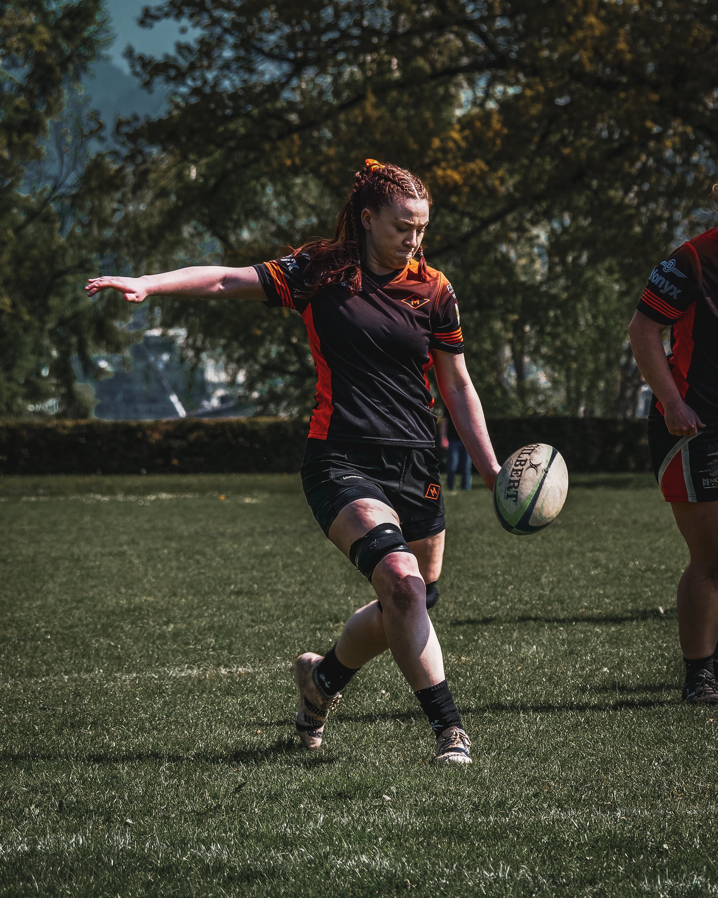 A female rugby player in black and red uniform kicking a rugby ball on a grassy field, with trees and blue sky in the background.
