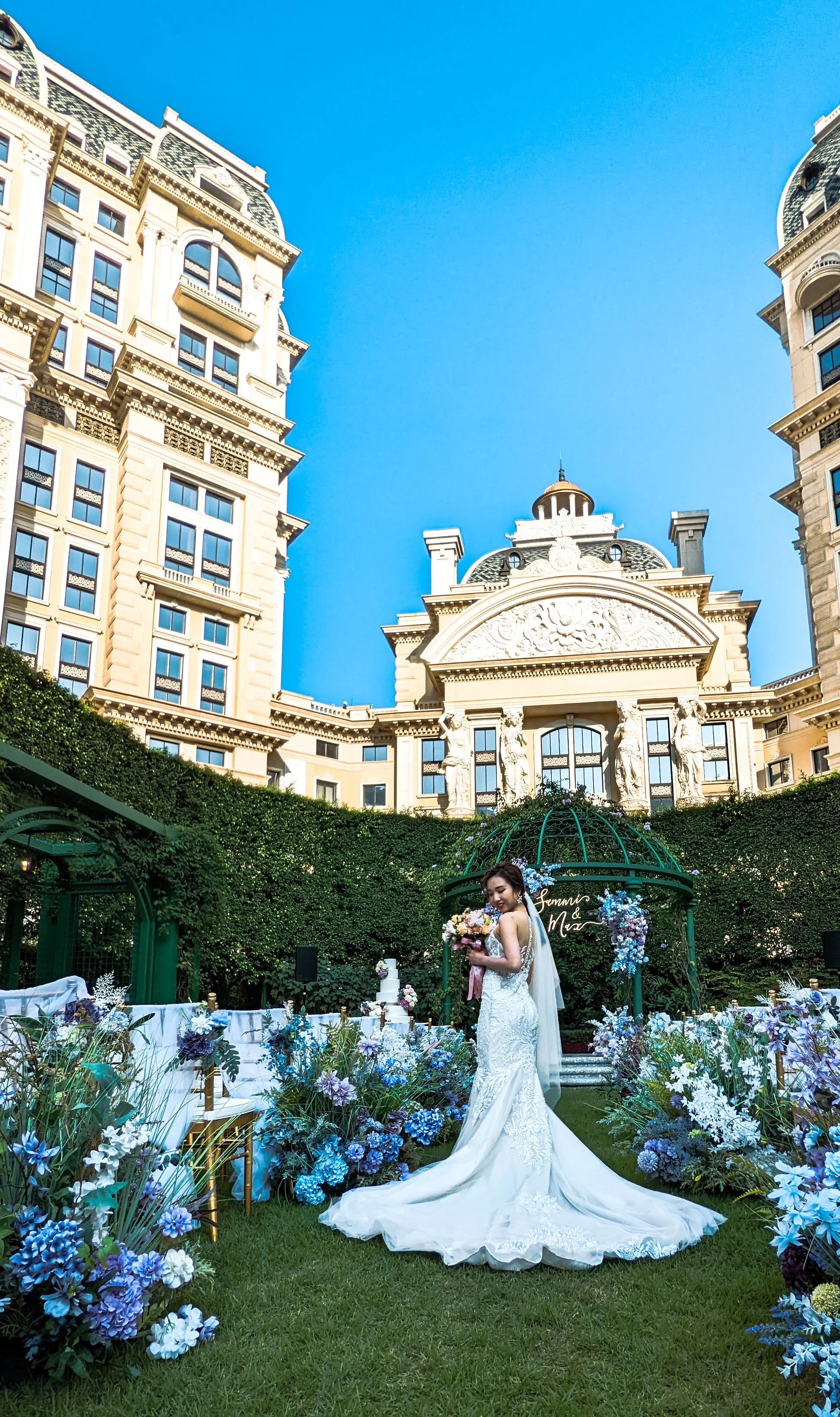 Bride in a wedding dress holding a bouquet, standing in an outdoor wedding setup with floral arrangements, surrounded by tall historic buildings under a clear blue sky.