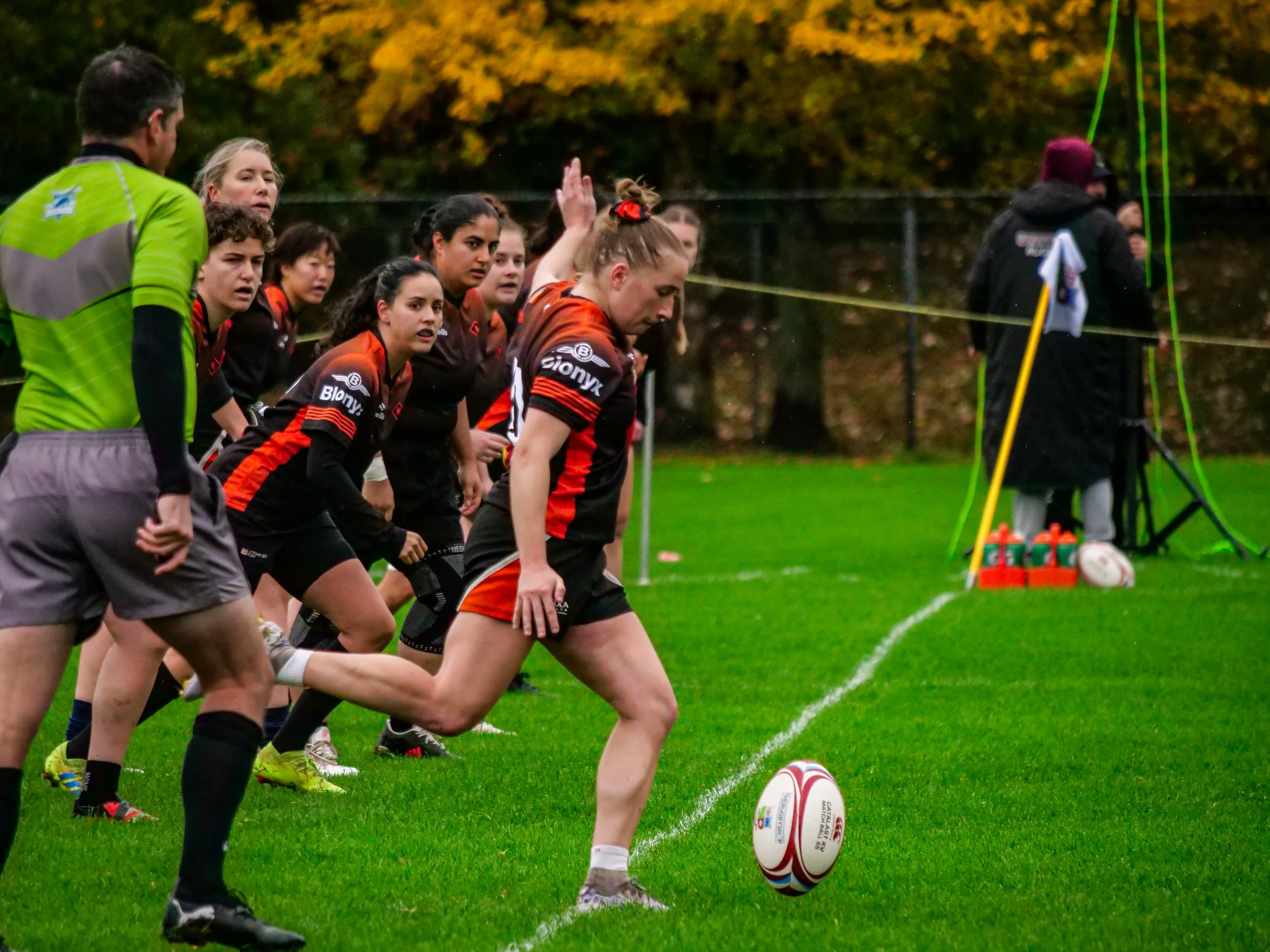 Women rugby team lining up for a kick on a grassy field during daytime with autumn trees in background