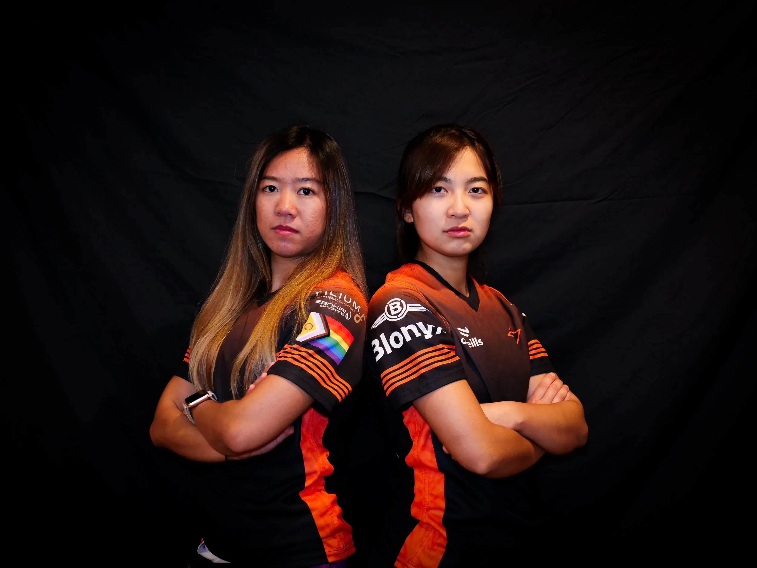 Two women with arms crossed, wearing black and orange sports jerseys, standing back to back against a black backdrop.